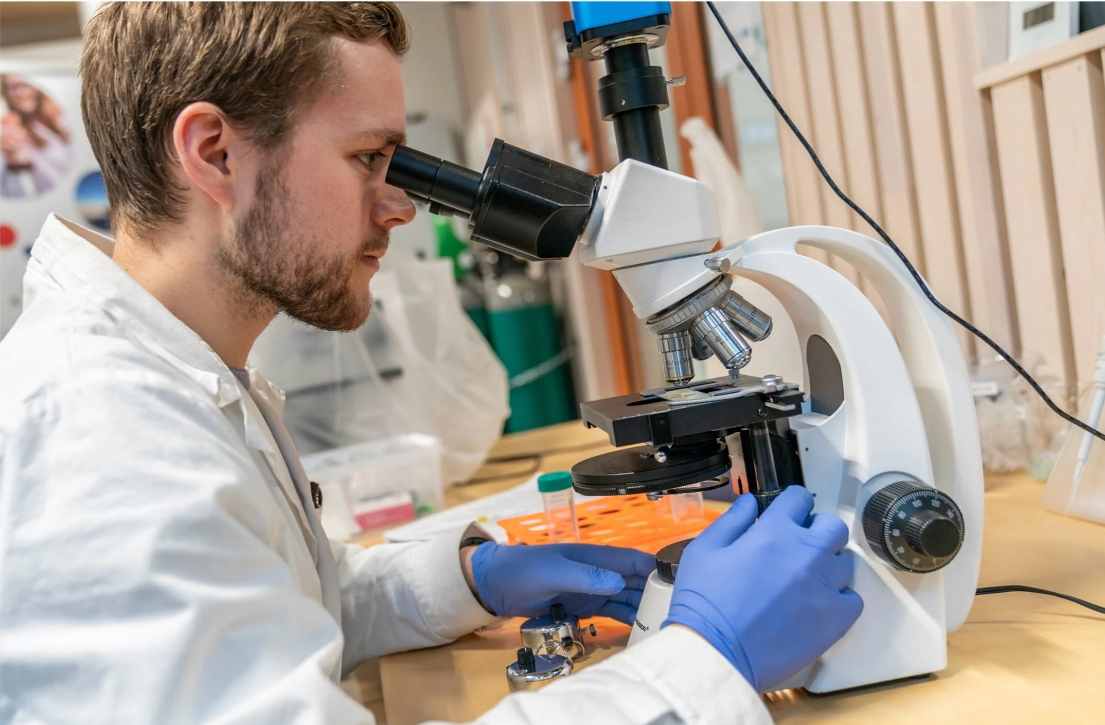 En person i hvit labfrakk ser gjennom et mikroskop i et laboratorium.