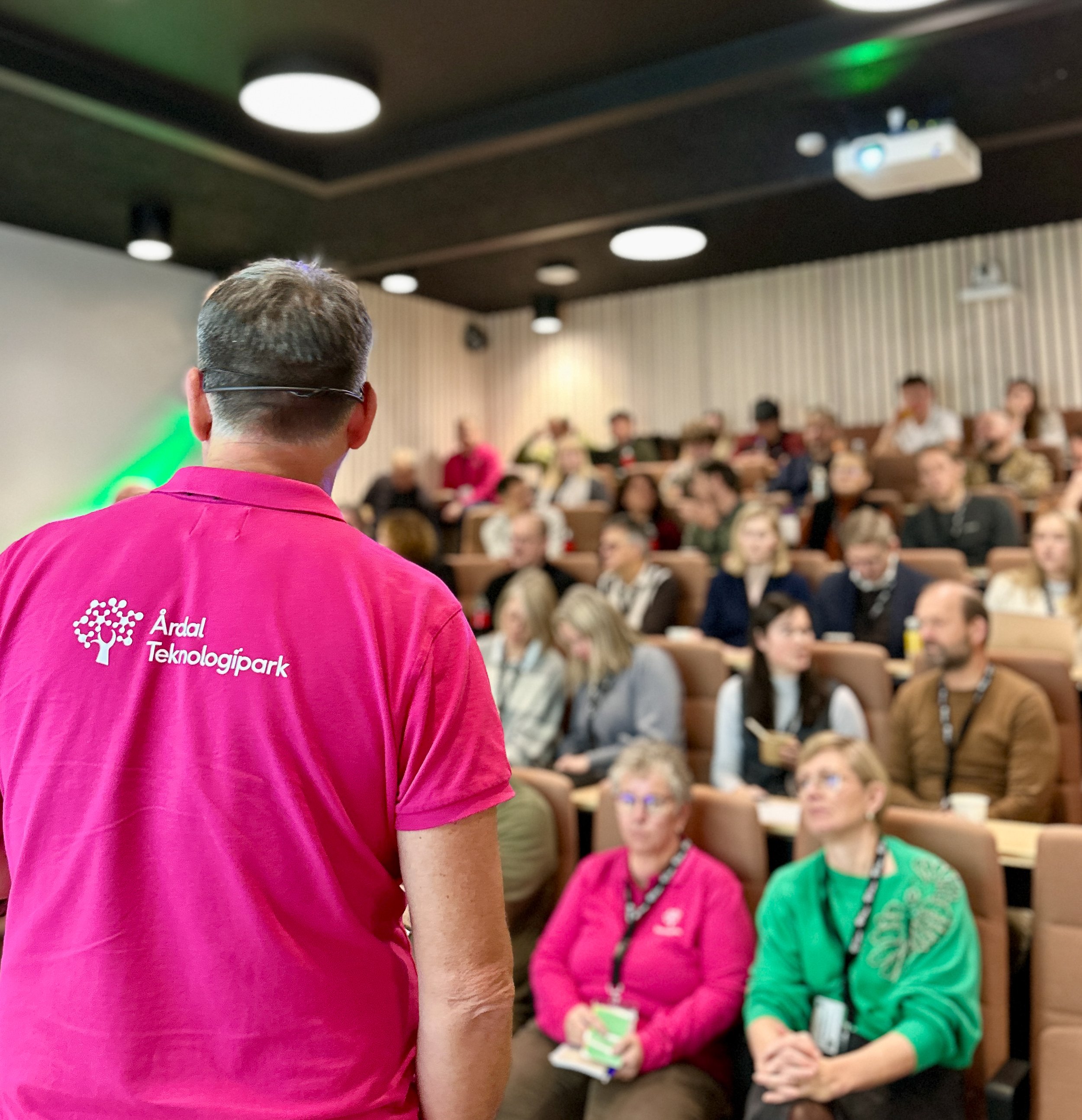 Person med rosa t-skjorte som har teksten «Årdal Teknologi» står foran et publikum i et auditorium, hvor folk sitter og lytter. Publikum består av en blanding av voksne og yngre personer, og noen tar notater.