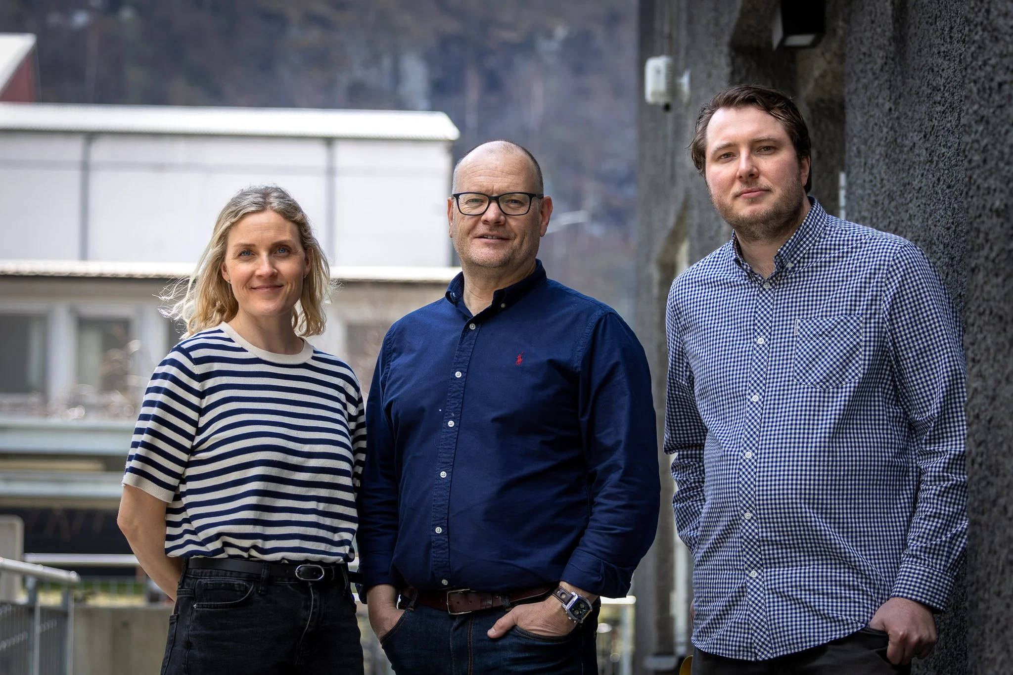 Tre personer står utendørs i en by eller boligområde, med bygninger og balkonger i bakgrunnen. De ser direkte mot kameraet og smiler.