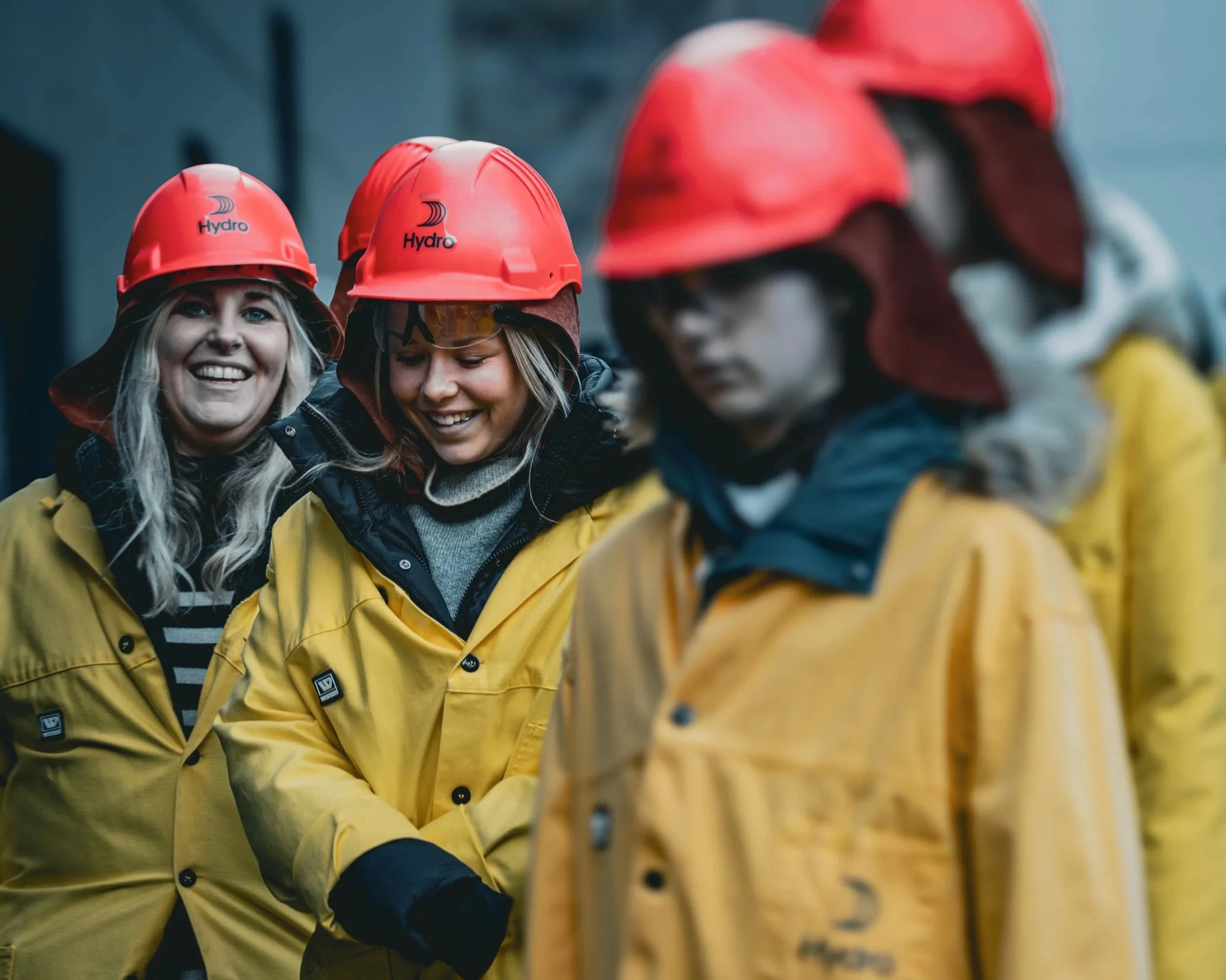To glade kvinner med lyse hår og smil, iført gule arbeidsjakker og røde hjelmer med logo, står i en gruppe utendørs.