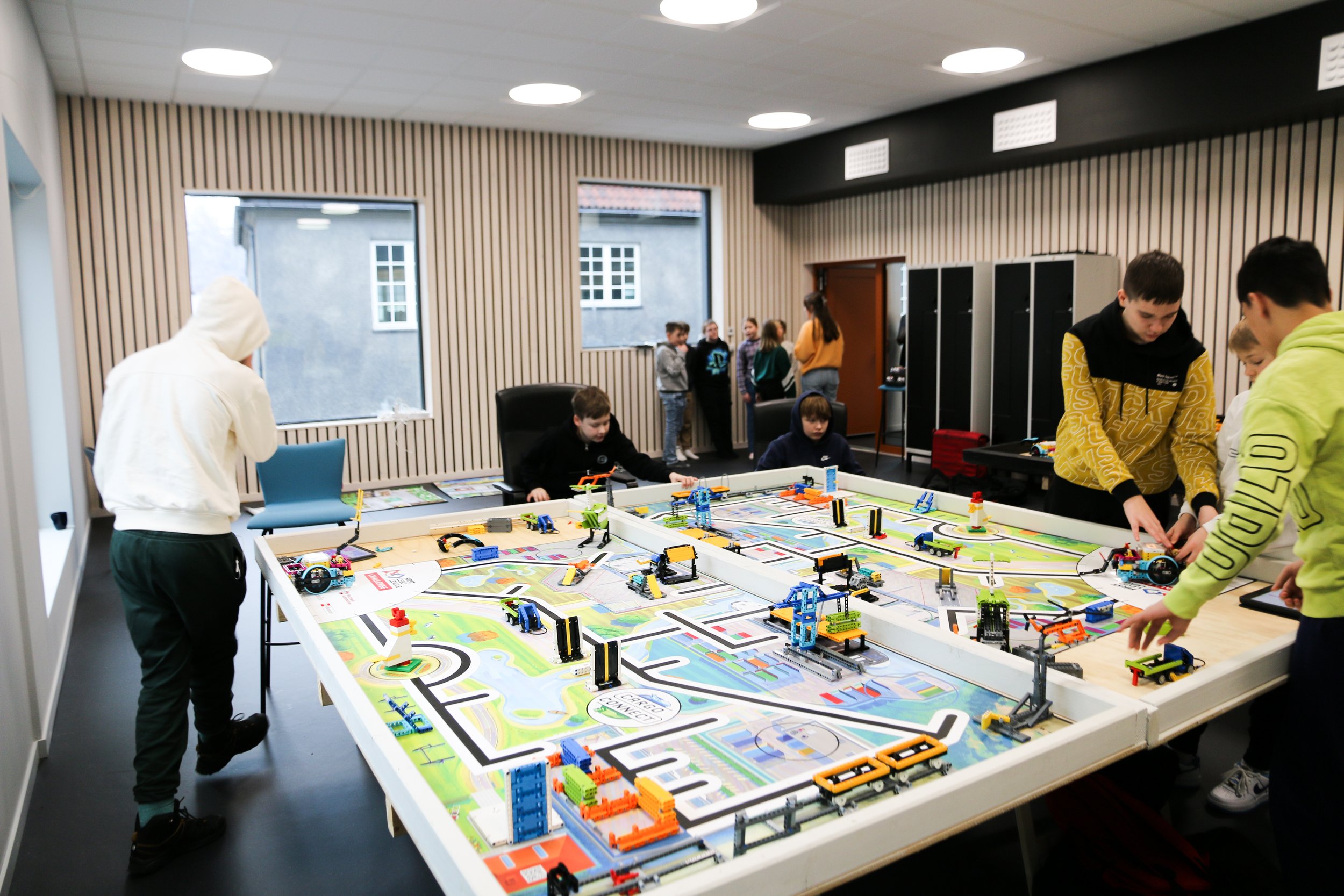 Barn og voksne som leker med LEGO-robotter på et bord i et rom med trestore vinduer og tre plafondlys.