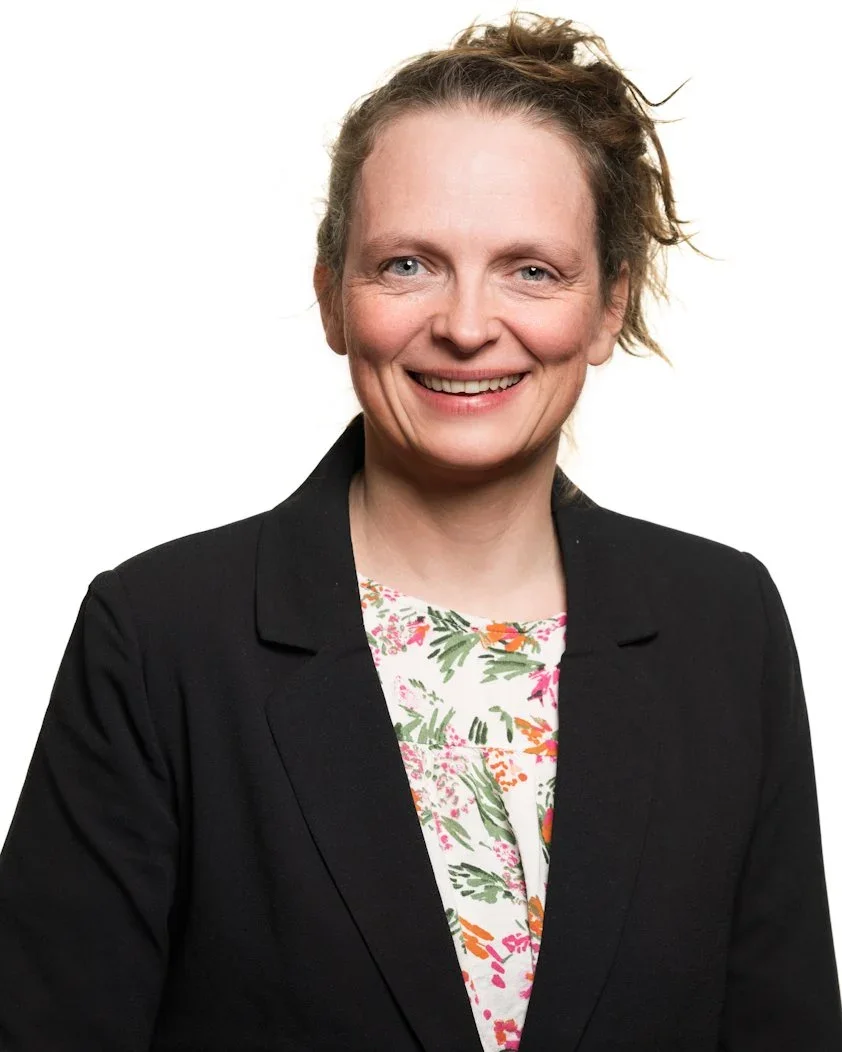 Et portrett av en kvinne med lys hud, lyst brunt hår og blå øyne, iført en svart jakke og en fargerik bluse, smilende mot kameraet.