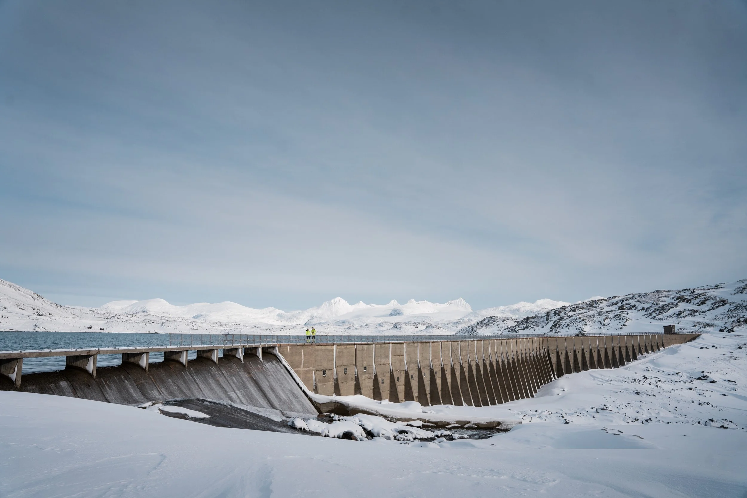 To personer står på en demning omgitt av snødekte fjell og vann, under en skyet himmel.
