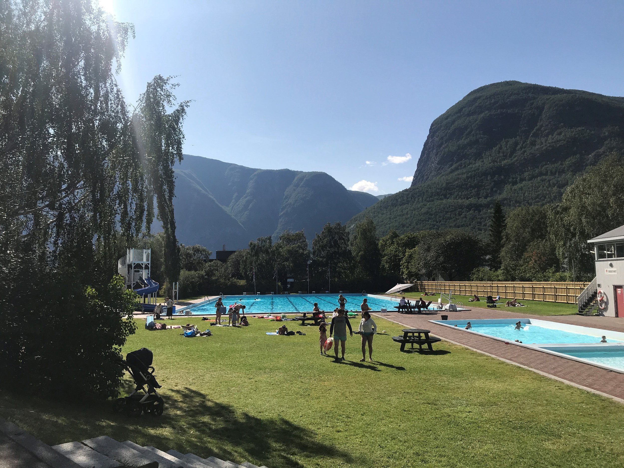 Barn og voksne bruker svømmebassenger i utendørs svømmebassengområde, omgitt av grønne trær og fjell i bakgrunnen på en solrik dag.