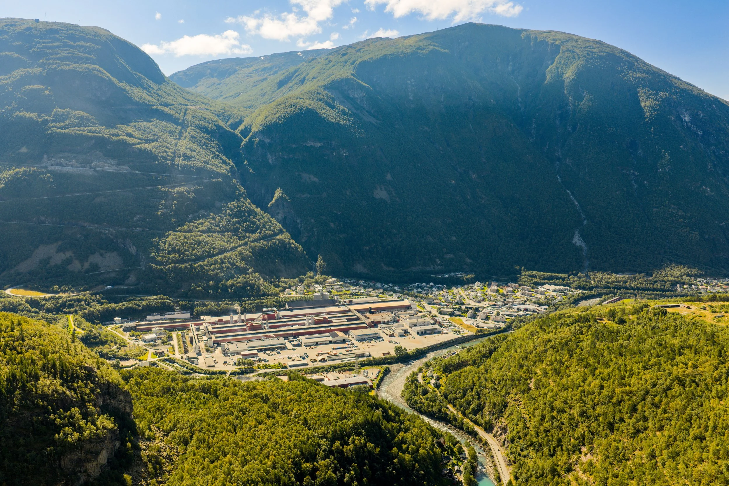 Bildet viser en by eller industriområde foran grønne fjell, med en elv som renner gjennom dalen.