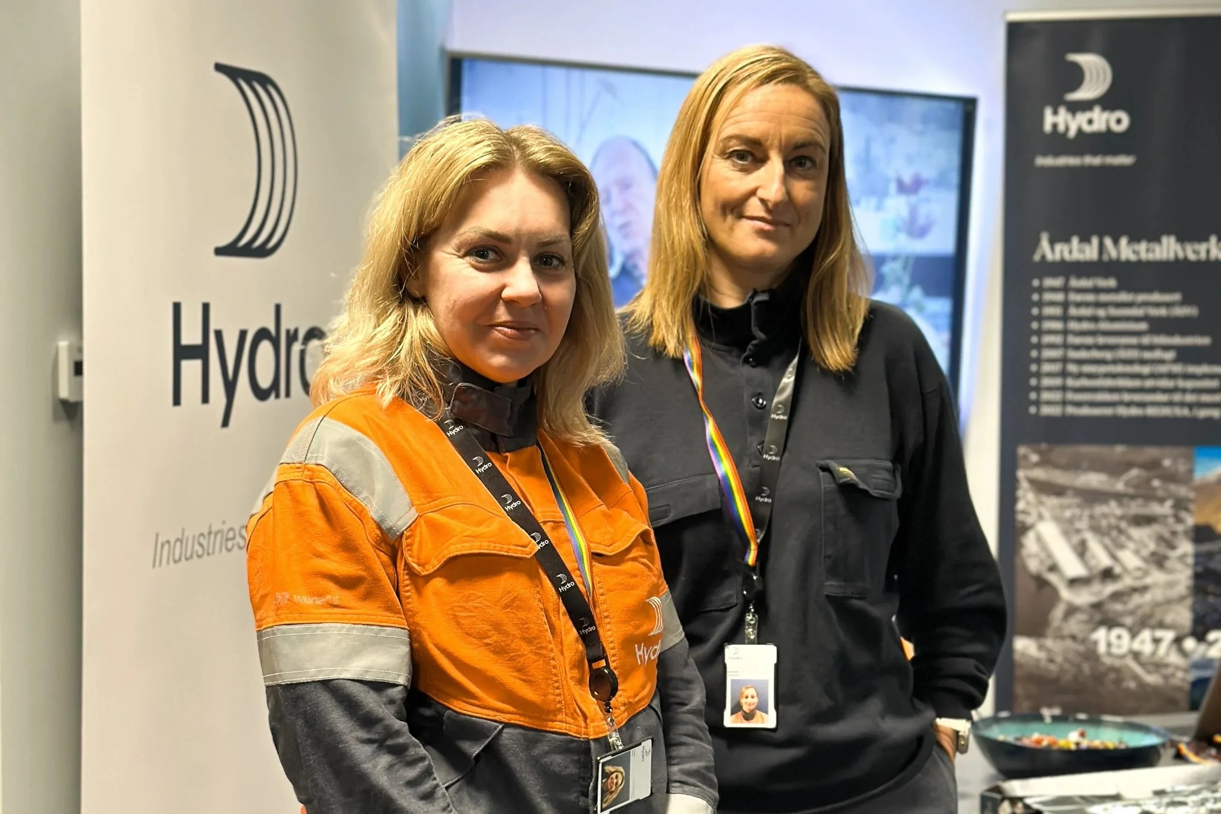 To kvinner står ved en messe eller utstilling, med ca. 1 meters mellomrom, iført jakker og med identitetskort rundt halsen. Bak dem er det en plakat med logoen Hydro og en annen plakat med tekst og bilder.
