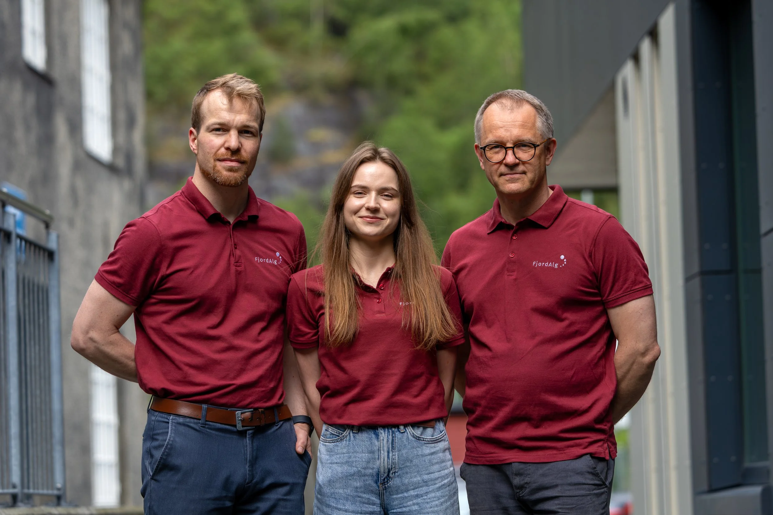 Tre personer står utendørs, alle iført burgunderrøde skjorter med logoet 'FjordAlg'.