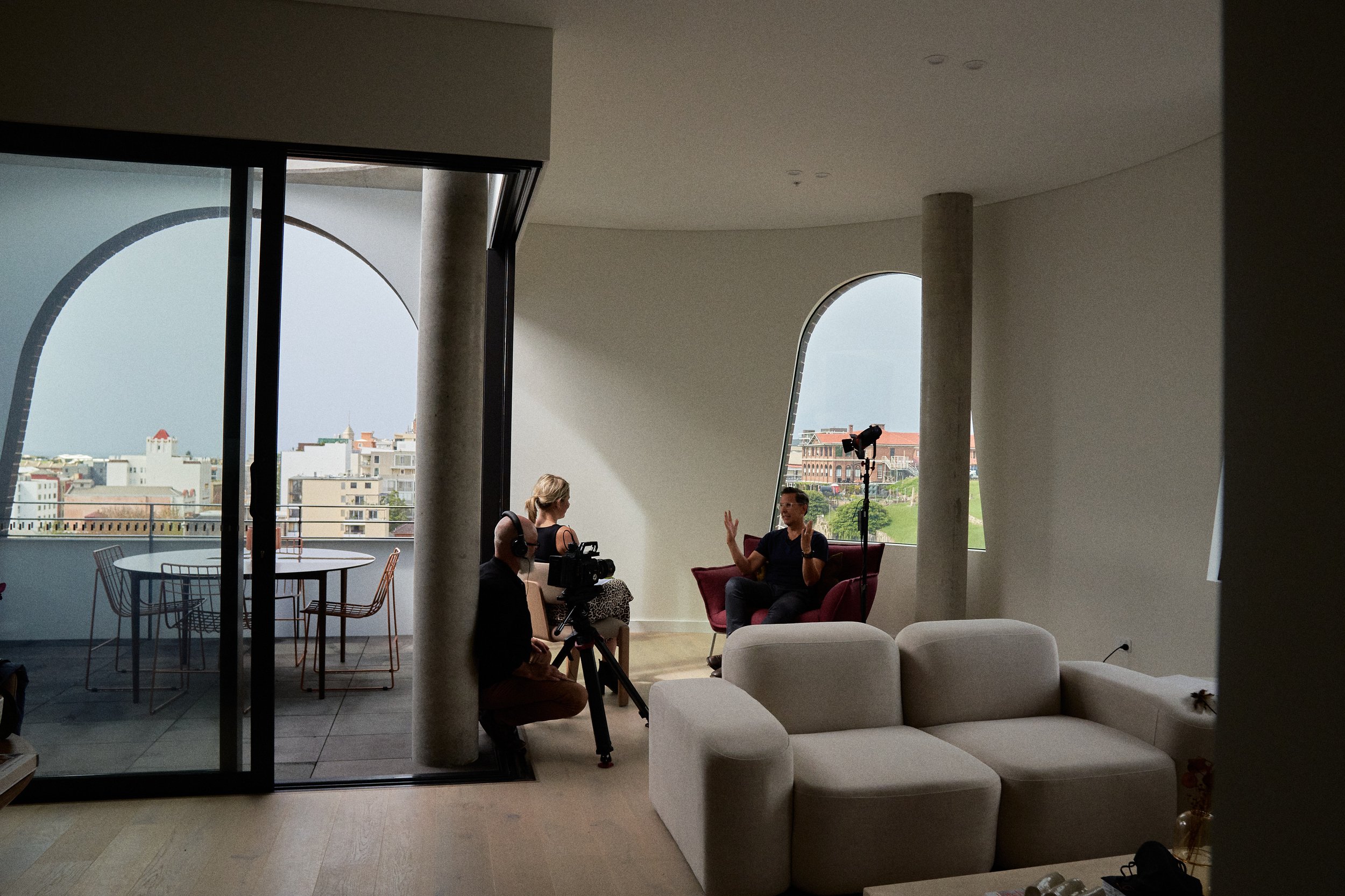 Modern living room with two people filming a seated man, large windows with city view, minimalist furniture.