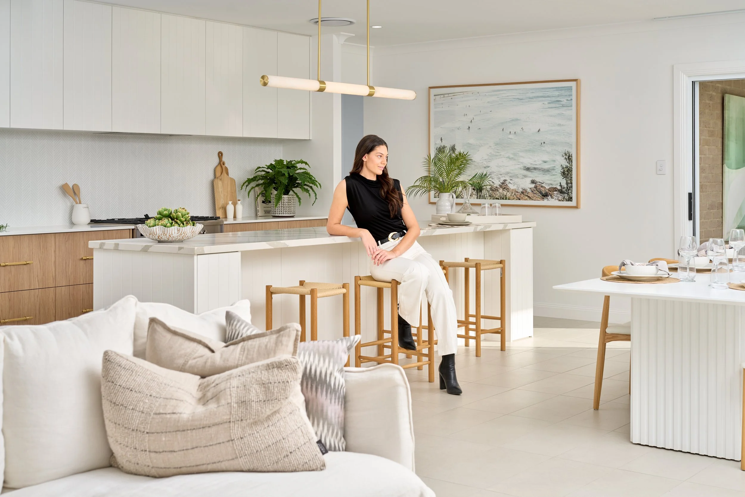 Woman sitting on a barstool at a kitchen island in a modern, bright kitchen with white cabinets, wooden accents, potted plants, and a large wall art of wave scene.