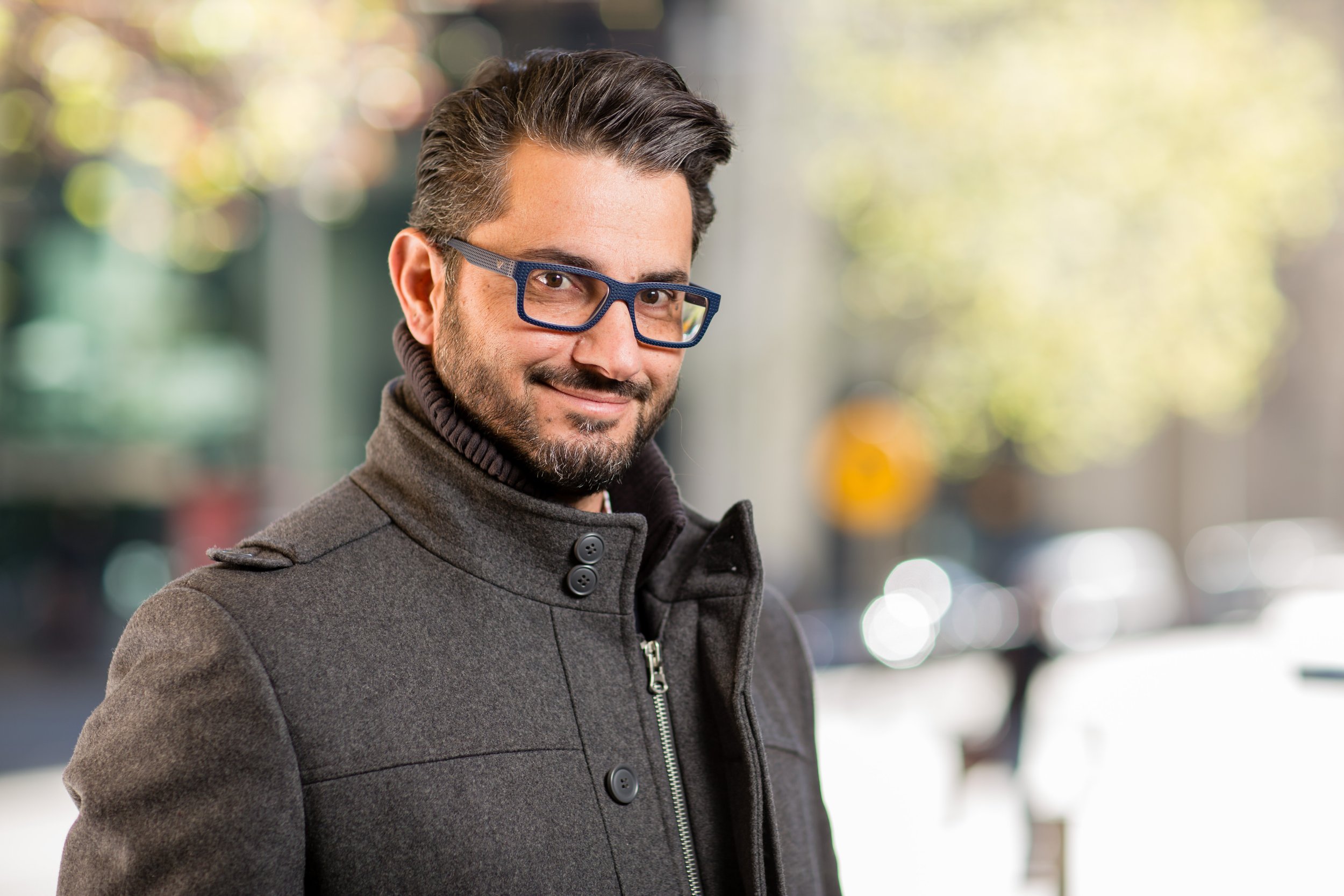 Man with glasses and a coat smiling outdoors with blurred background.