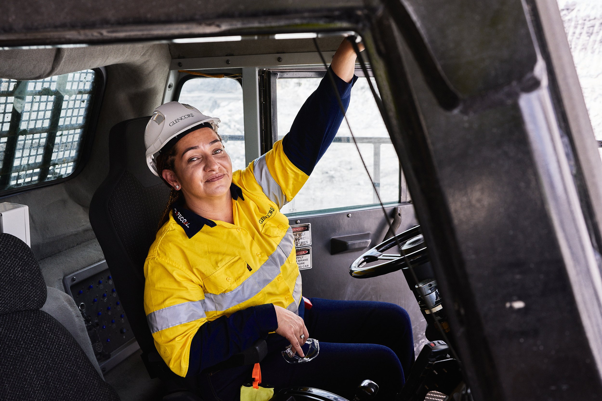 A person wearing a white hard hat and yellow high-visibility work jacket is sitting in the cab of a vehicle, possibly a construction or mining vehicle, with one hand on the steering wheel and the other adjusting controls. The person is looking at the