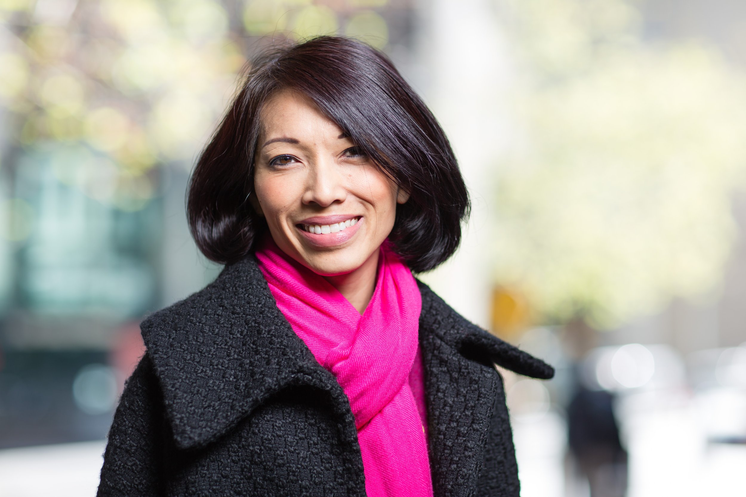 Smiling woman with short dark hair wearing black coat and pink scarf outdoors.
