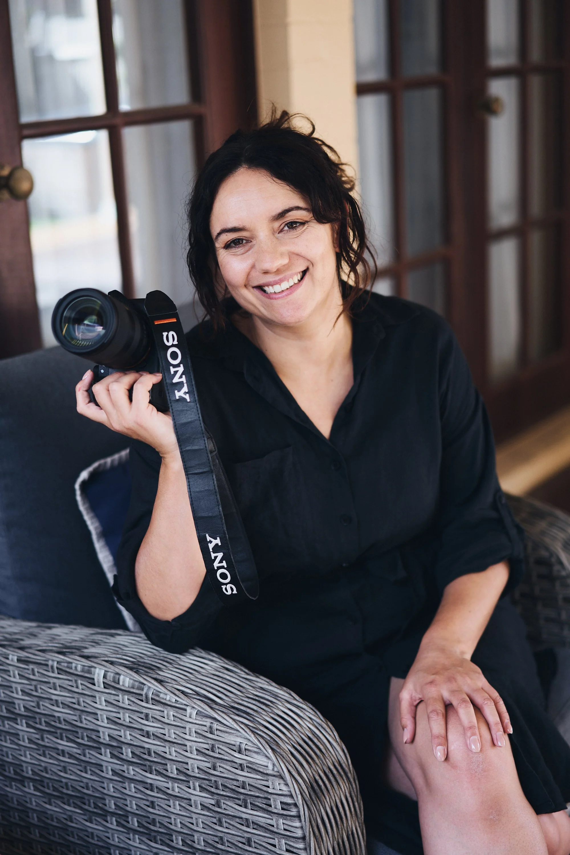 Smiling woman sitting on a wicker chair inside a room, holding a Sony camera over her shoulder.