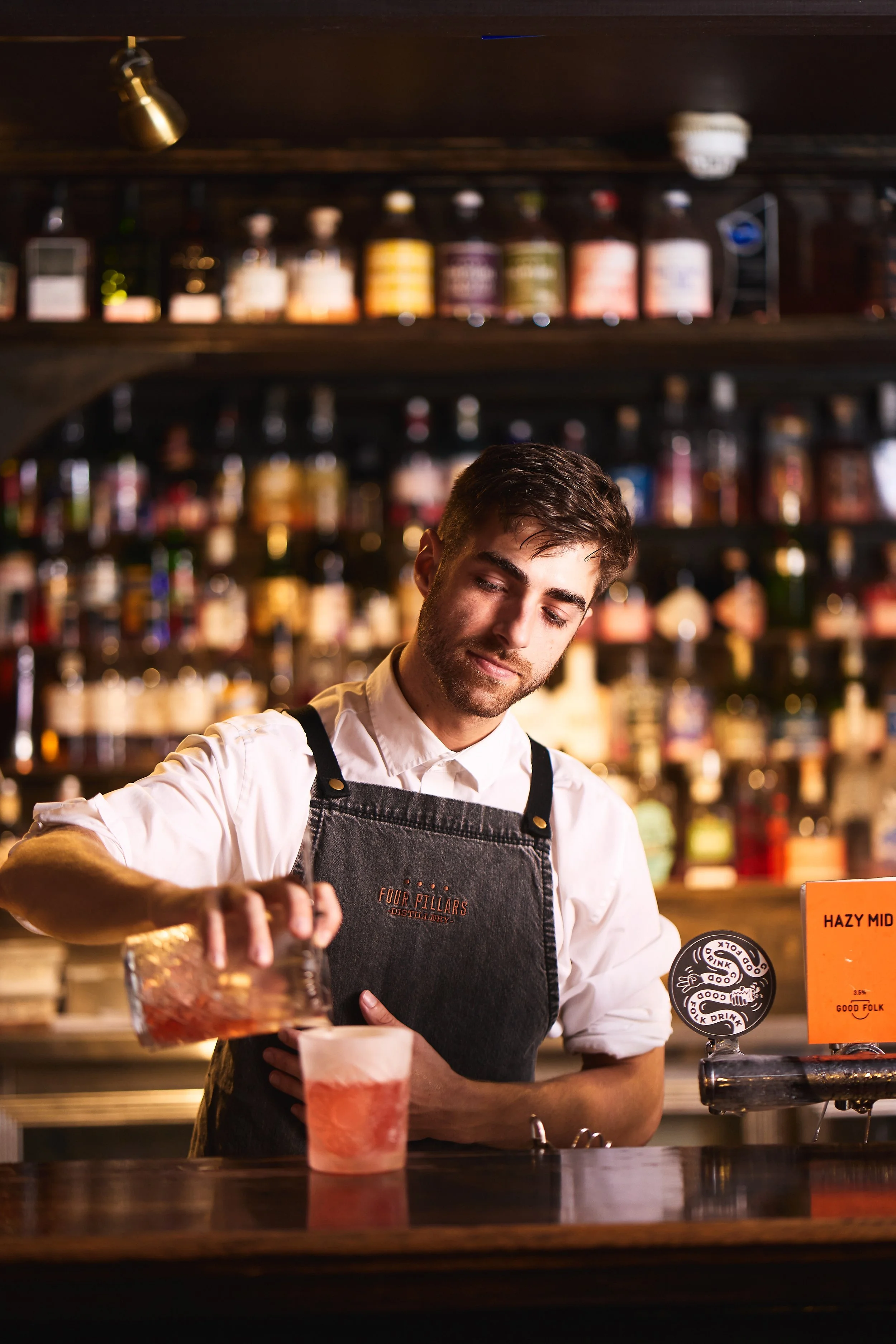 A bartender pouring a drink at a bar, with shelves of various bottles in the background.
