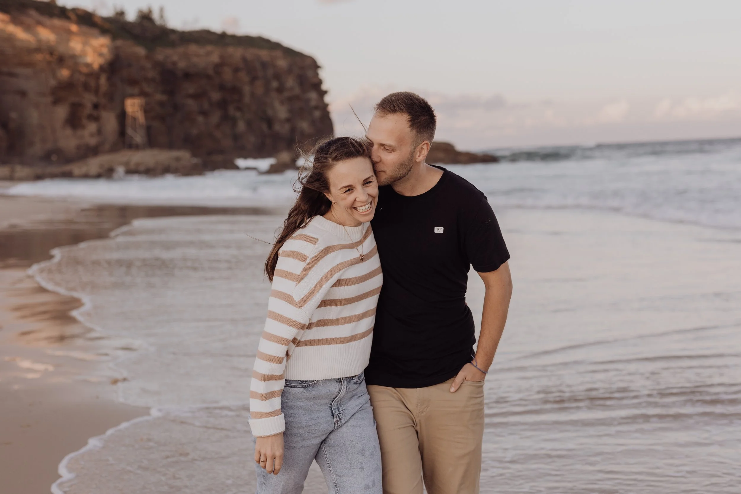 Couple smiling on a beach with cliffs and ocean in the background.