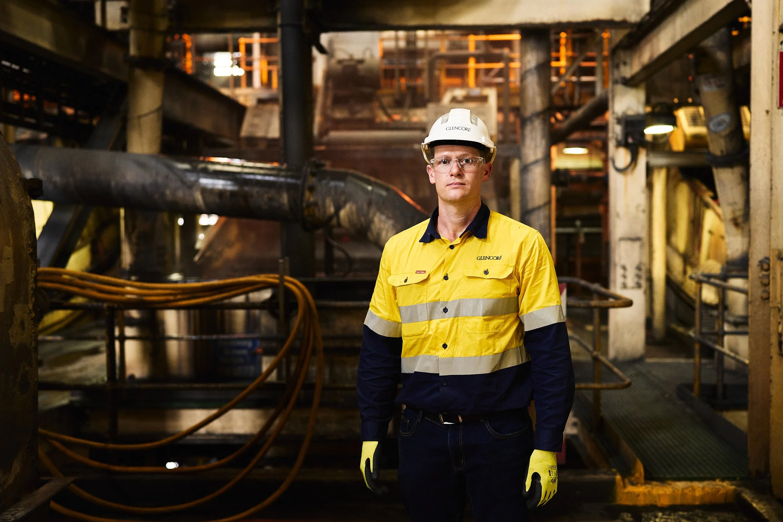 Man in yellow safety jacket and hard hat in industrial setting with machinery.