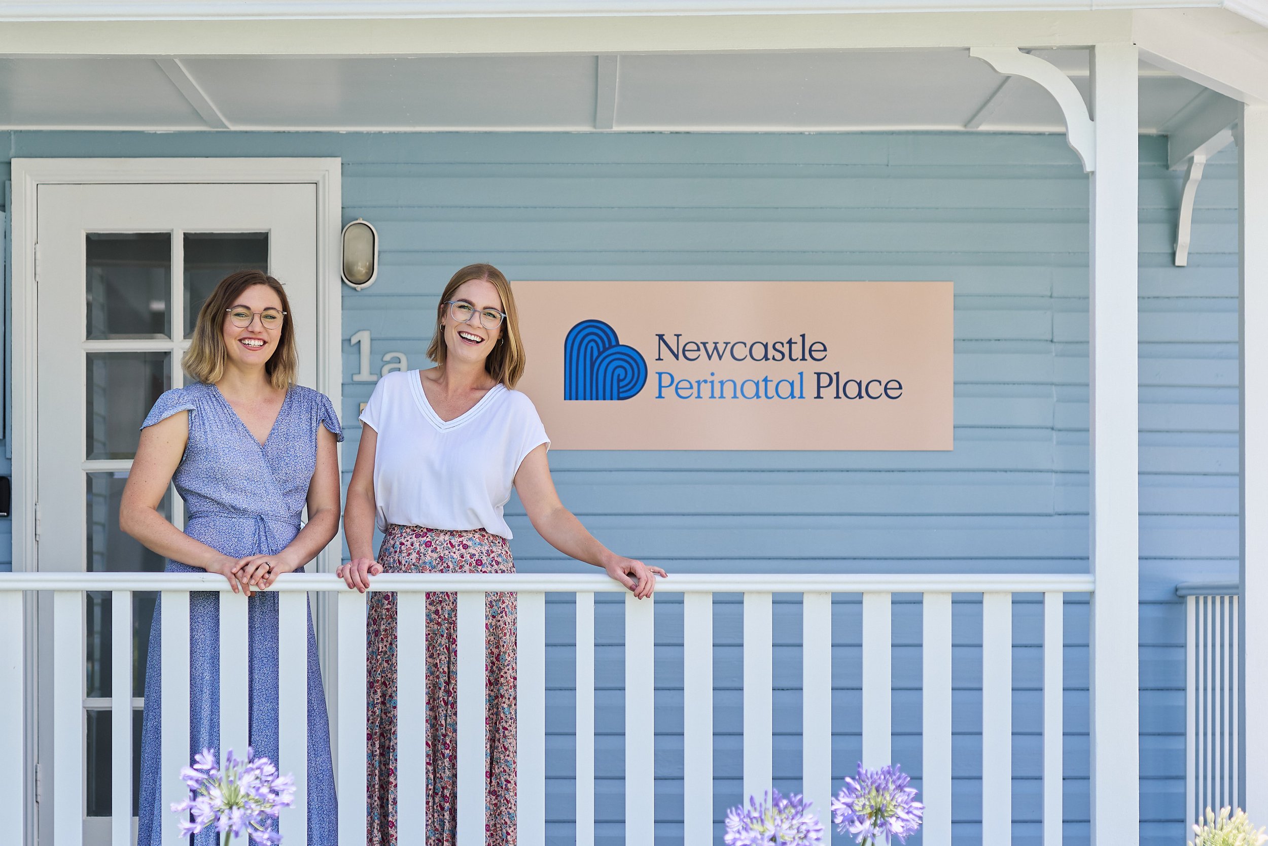 Two women standing on a porch of a light blue building with a sign reading "Newcastle Perinatal Place."
