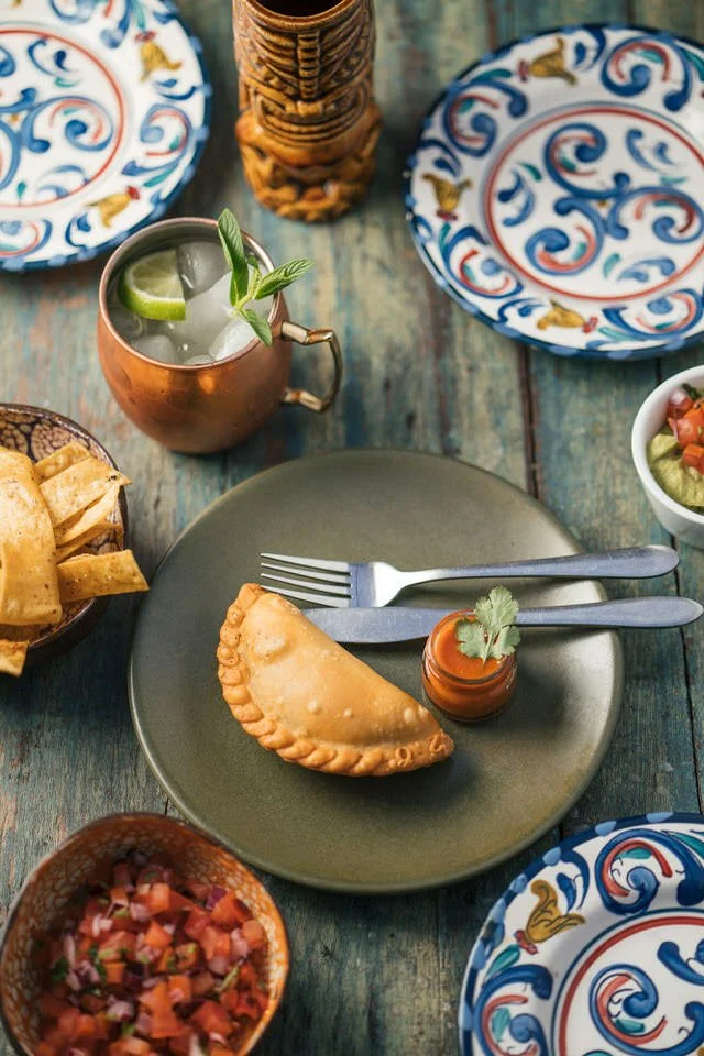 A table setting with a filled turnover on a plate, surrounded by various colorful plates and bowls containing toppings like salsa and guacamole. A copper mug with a drink and lime wedges is also present.