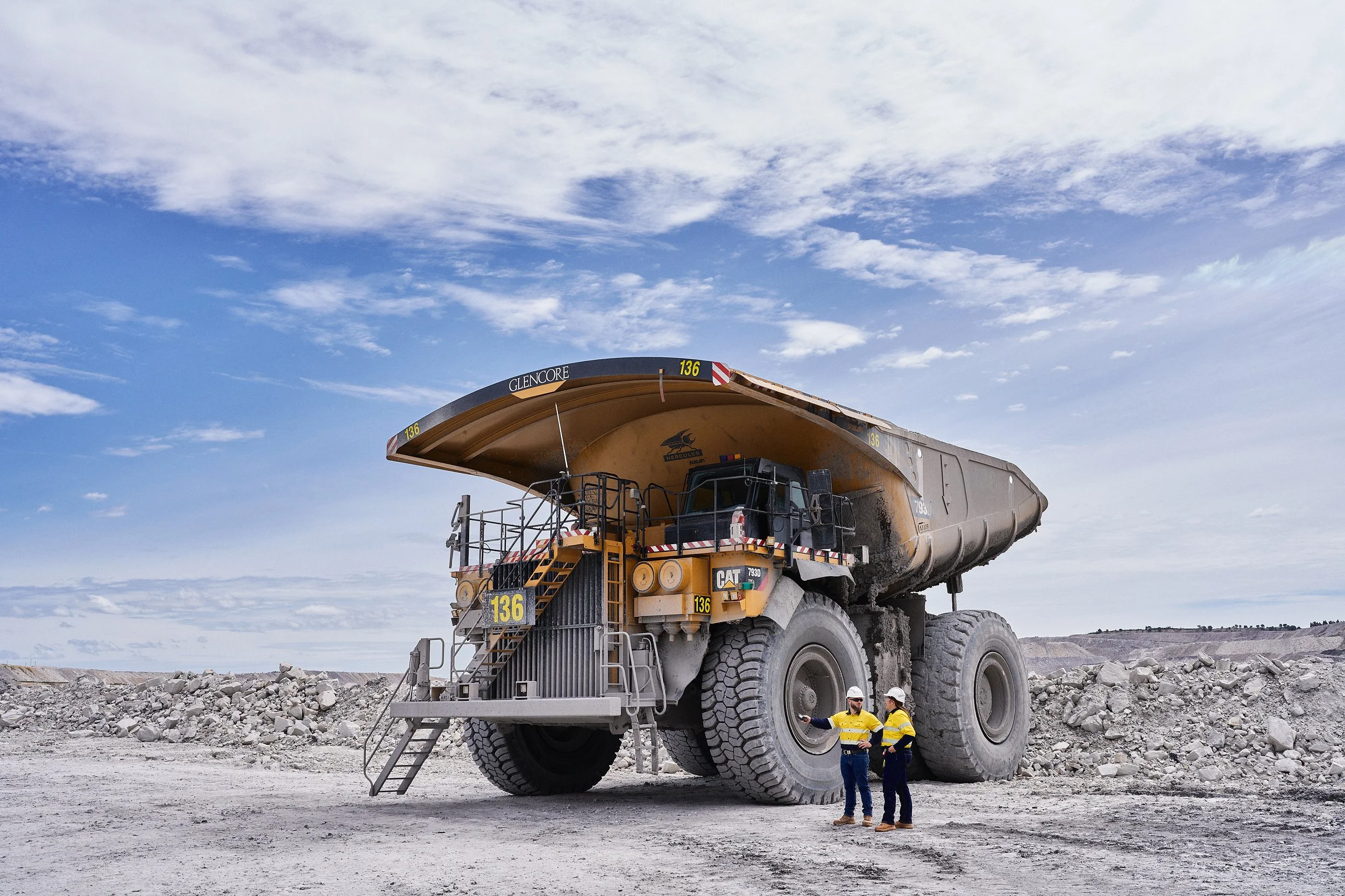 A large mining dump truck with two workers in safety gear standing nearby, under a blue sky.