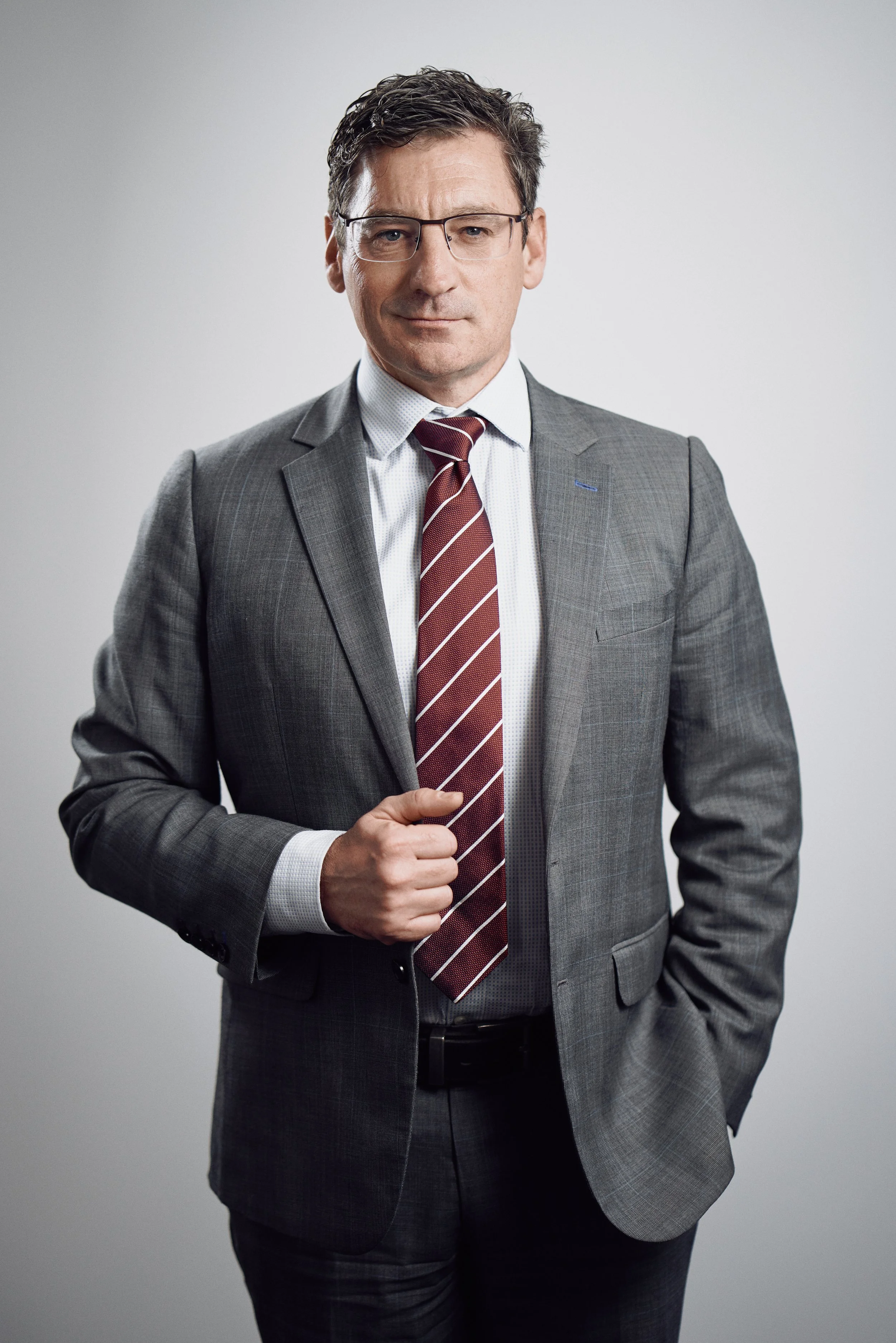 Man wearing a gray suit and red striped tie, standing in a confident pose against a neutral background.