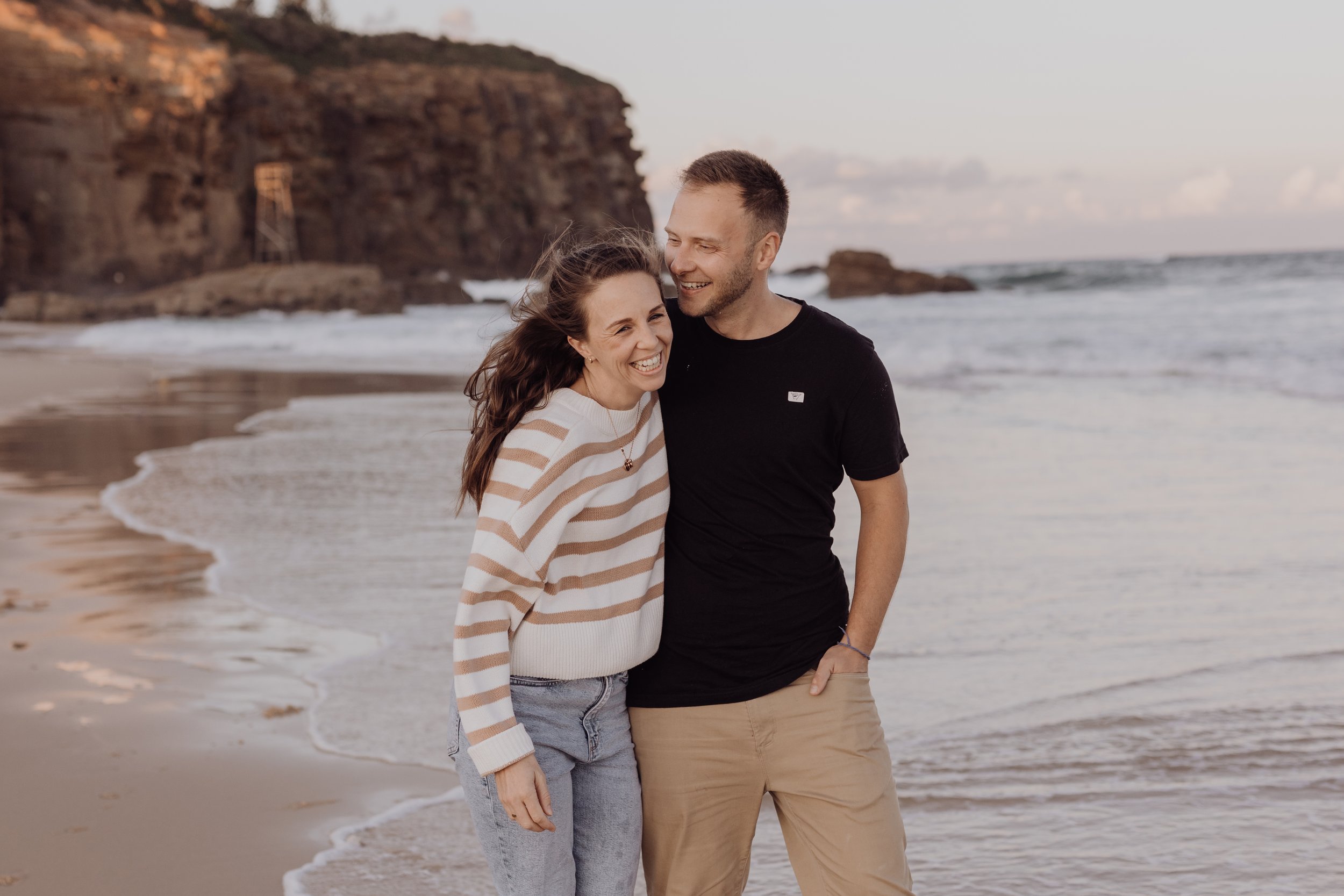 A smiling couple standing on a beach near the water, with rocky cliffs in the background during sunset.