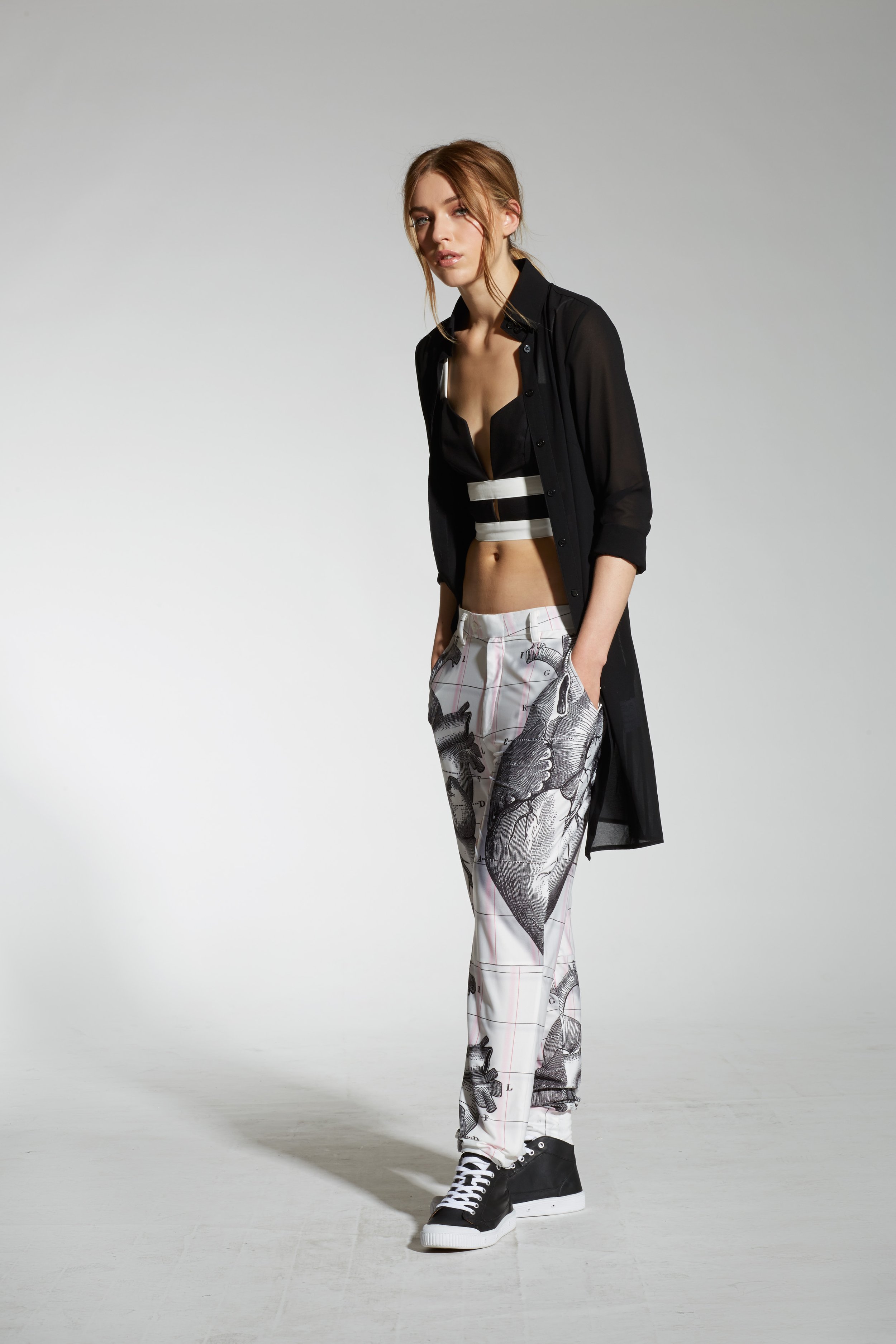 Fashionable woman wearing black crop top and long shirt, patterned pants, and sneakers against a plain background.
