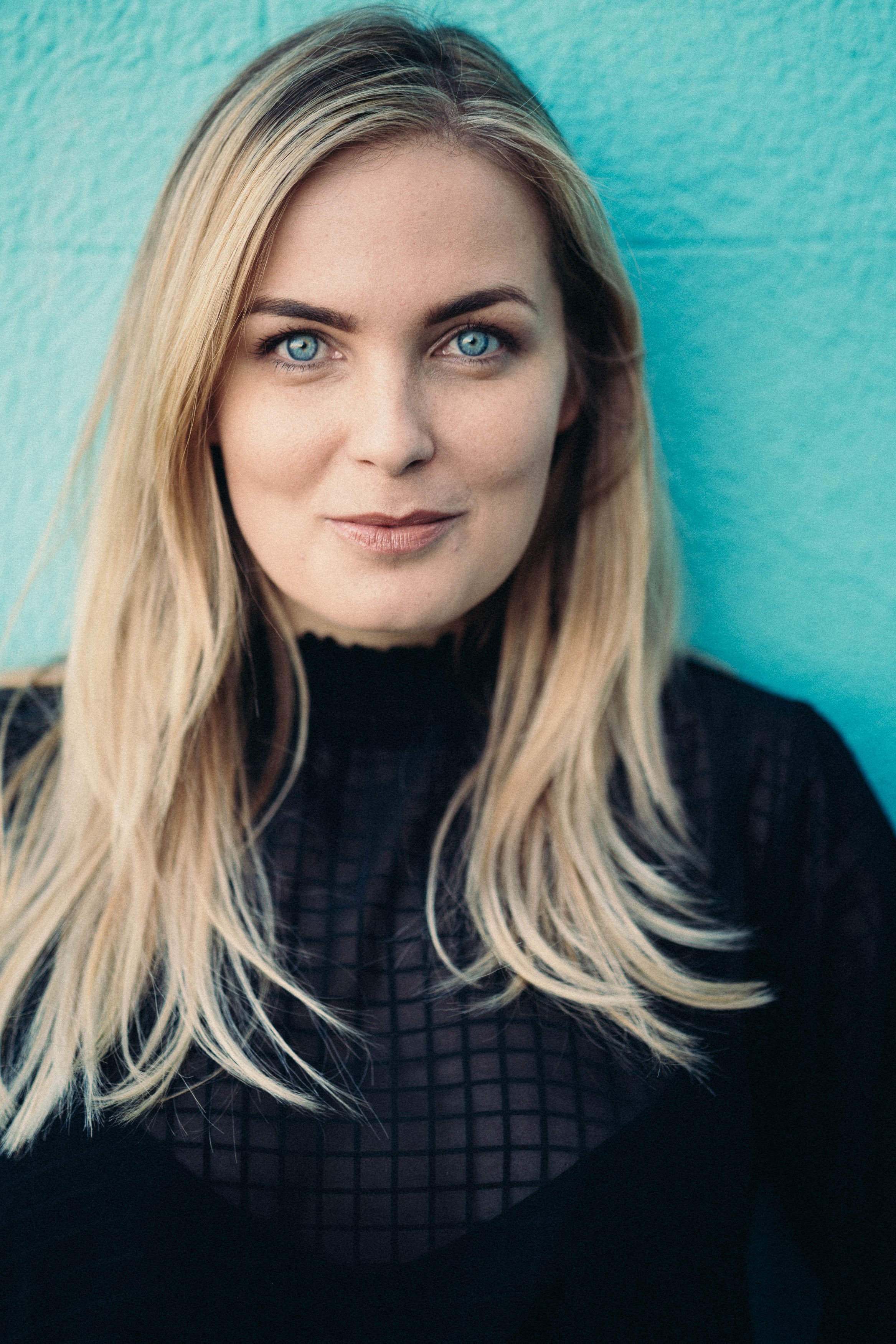 Woman with blonde hair and blue eyes, wearing a black patterned top, standing against a turquoise background.