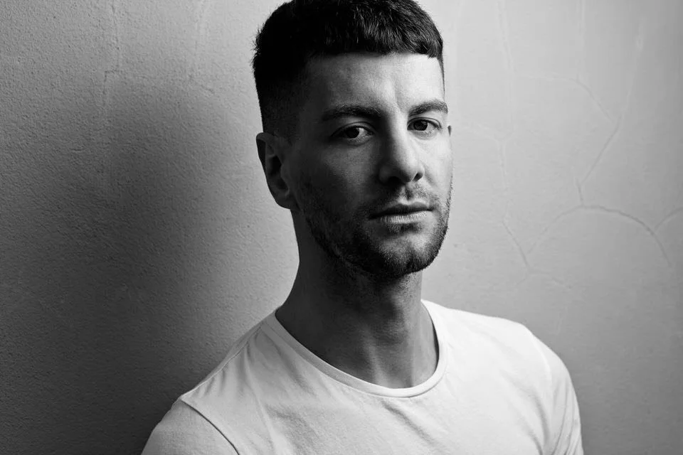 Black and white portrait of a man wearing a white t-shirt, with short hair, against a textured wall.