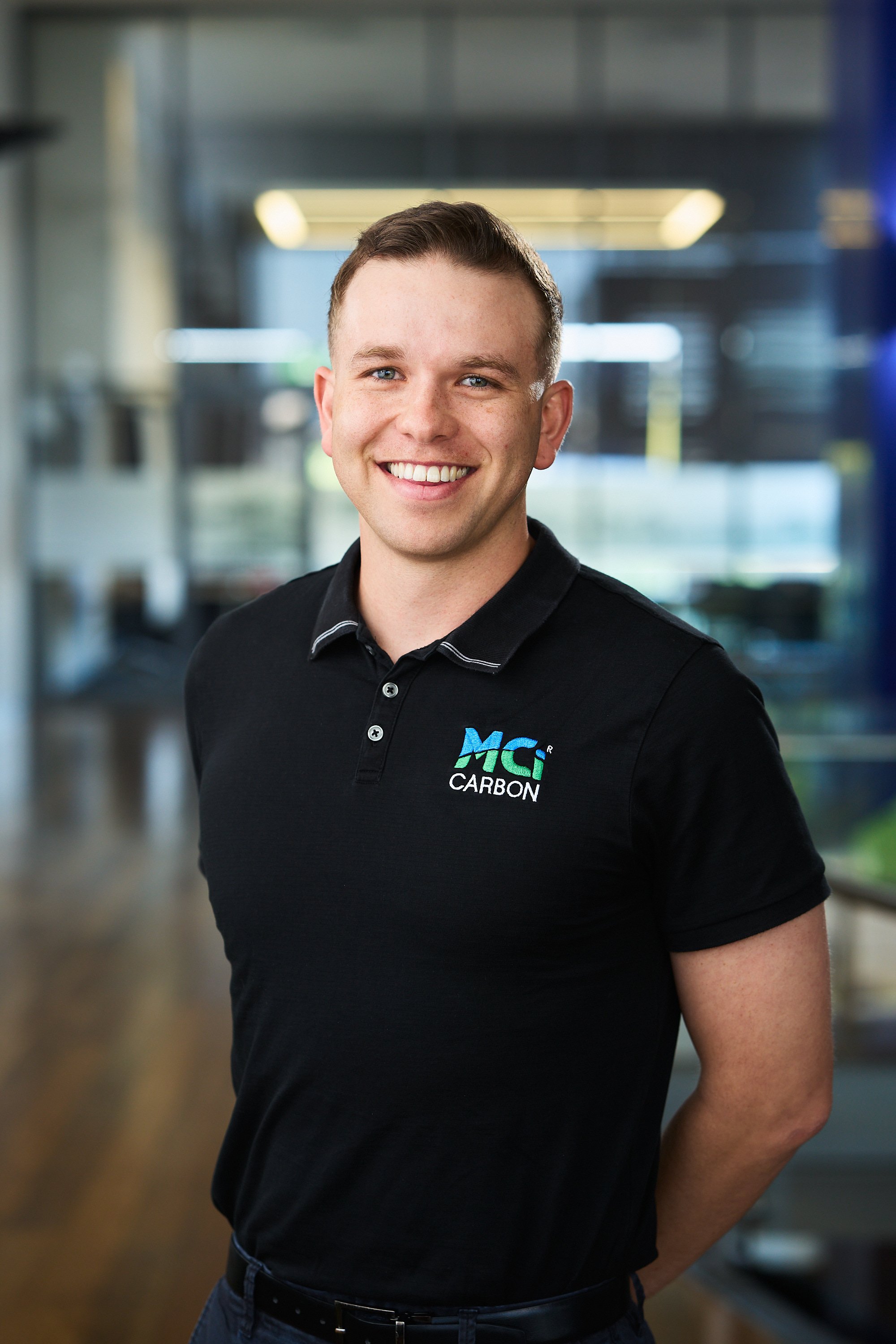 Person smiling in a black MCI Carbon polo shirt, standing in an office setting.