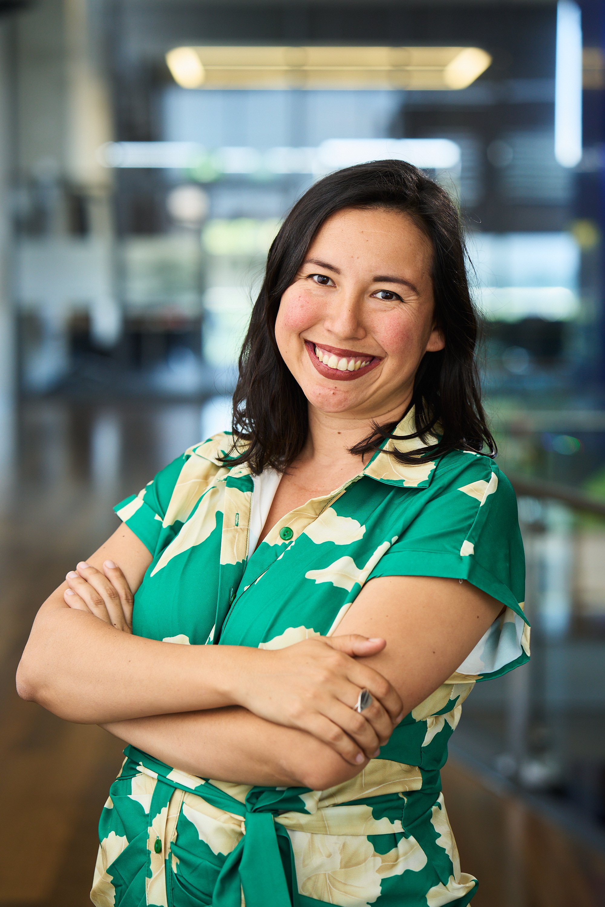 Smiling woman in a green floral dress with arms crossed, standing in an office environment.