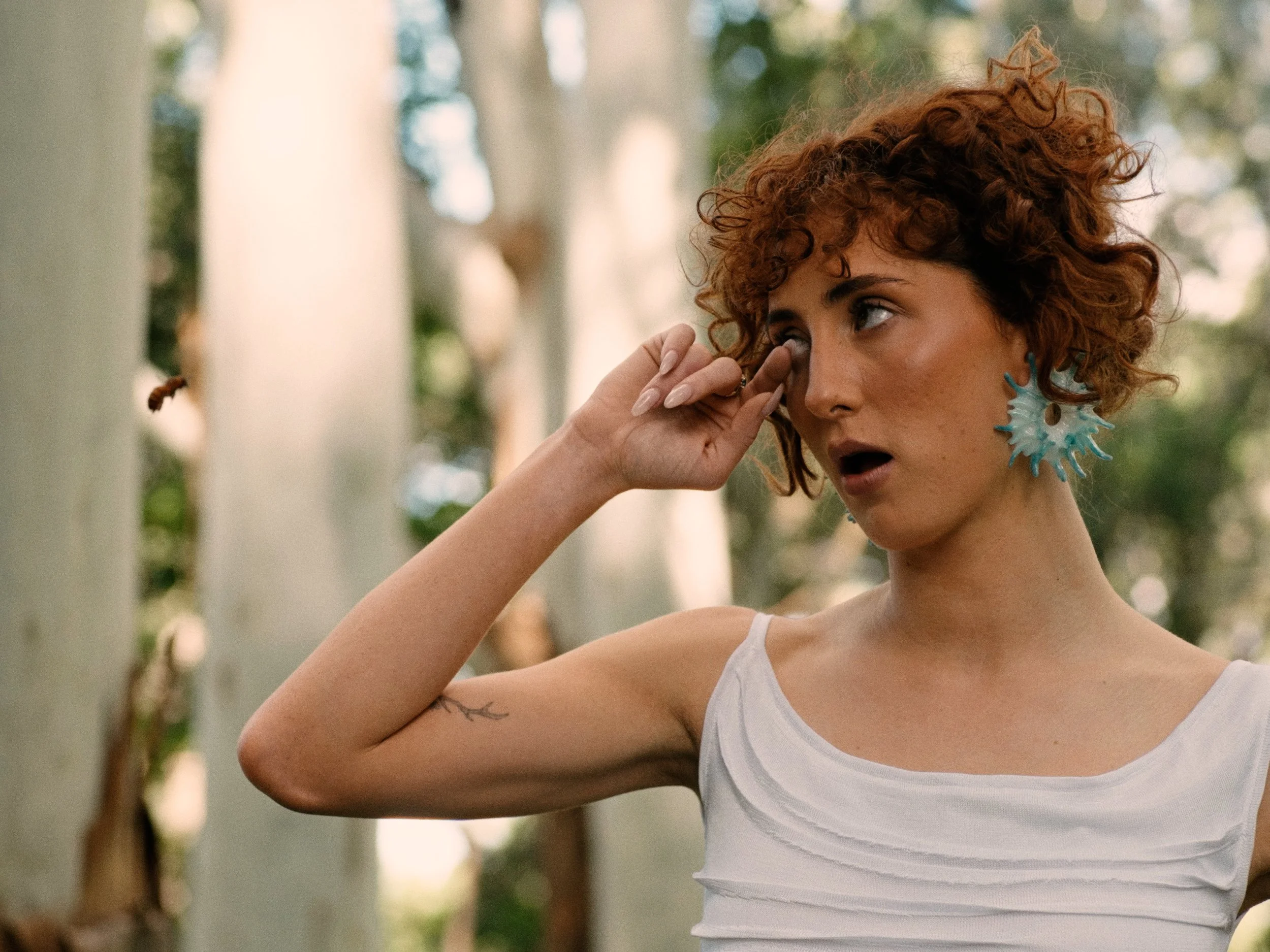 A woman with curly reddish hair and large blue earrings looks to the side while touching her eye, standing outdoors with trees in the background.