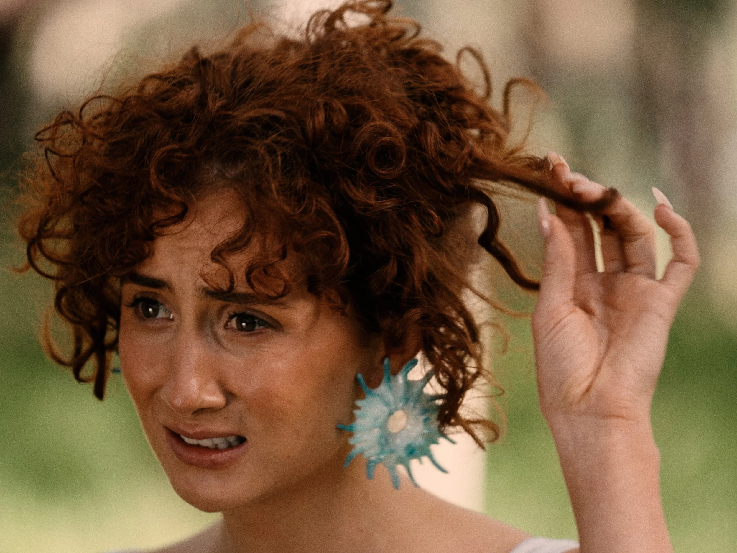 A woman with curly red hair wearing earrings and an off-shoulder top, smiling and holding a blue snowflake-shaped ornament near her head.