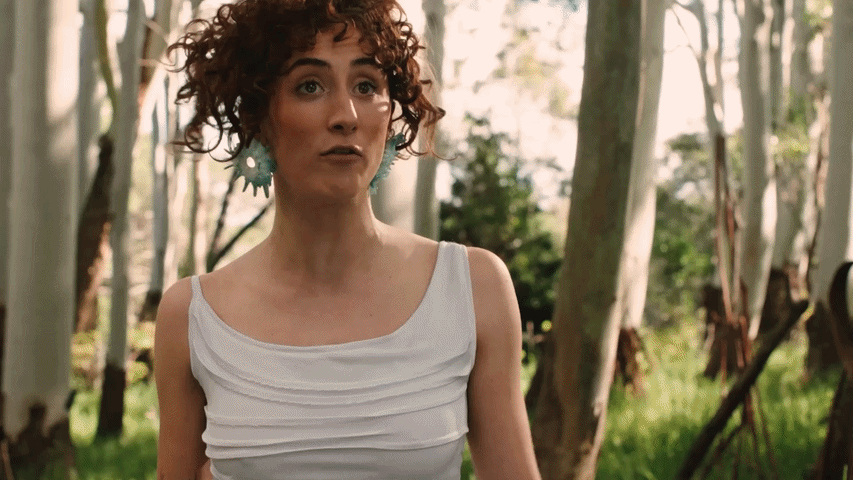 A woman with curly hair wearing a white top and large earrings standing outdoors among trees.