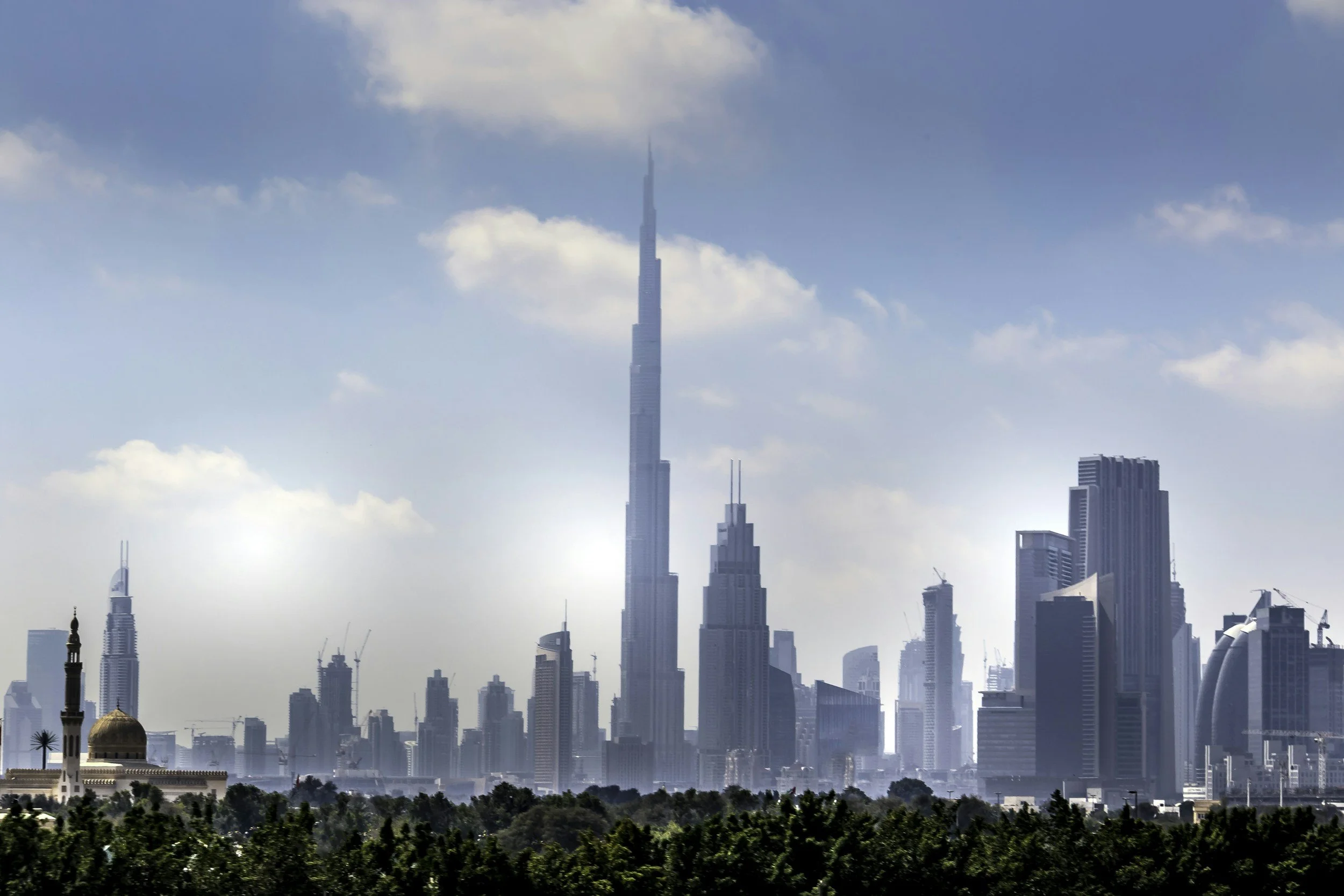 City skyline featuring the Burj Khalifa towering above other skyscrapers, with a mosque and greenery in the foreground, under a partly cloudy sky.