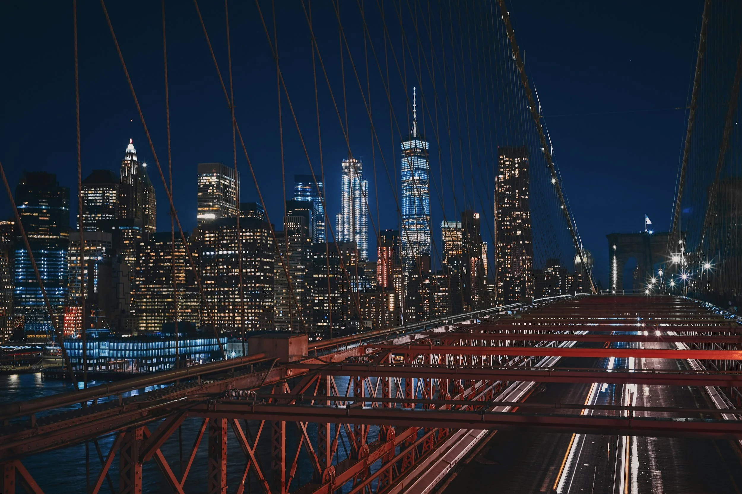 Nighttime view of Manhattan skyline from Brooklyn Bridge with illuminated skyscrapers, including the Empire State Building and One World Trade Center, and the bridge's red steel cables and structure in the foreground.