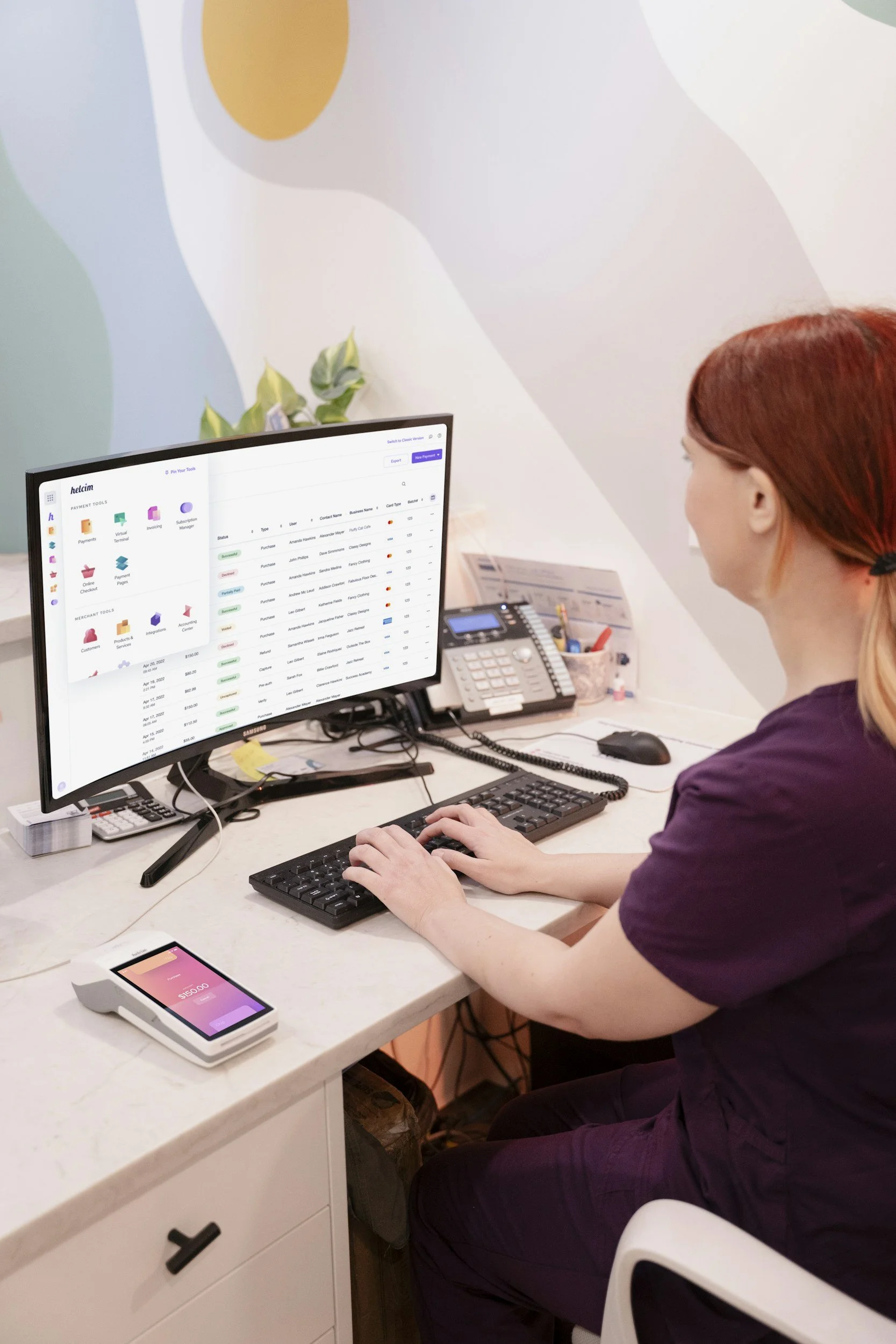 A woman with red hair working on a computer at an office desk, with a calculator, a smartphone, and office supplies on the desk.
