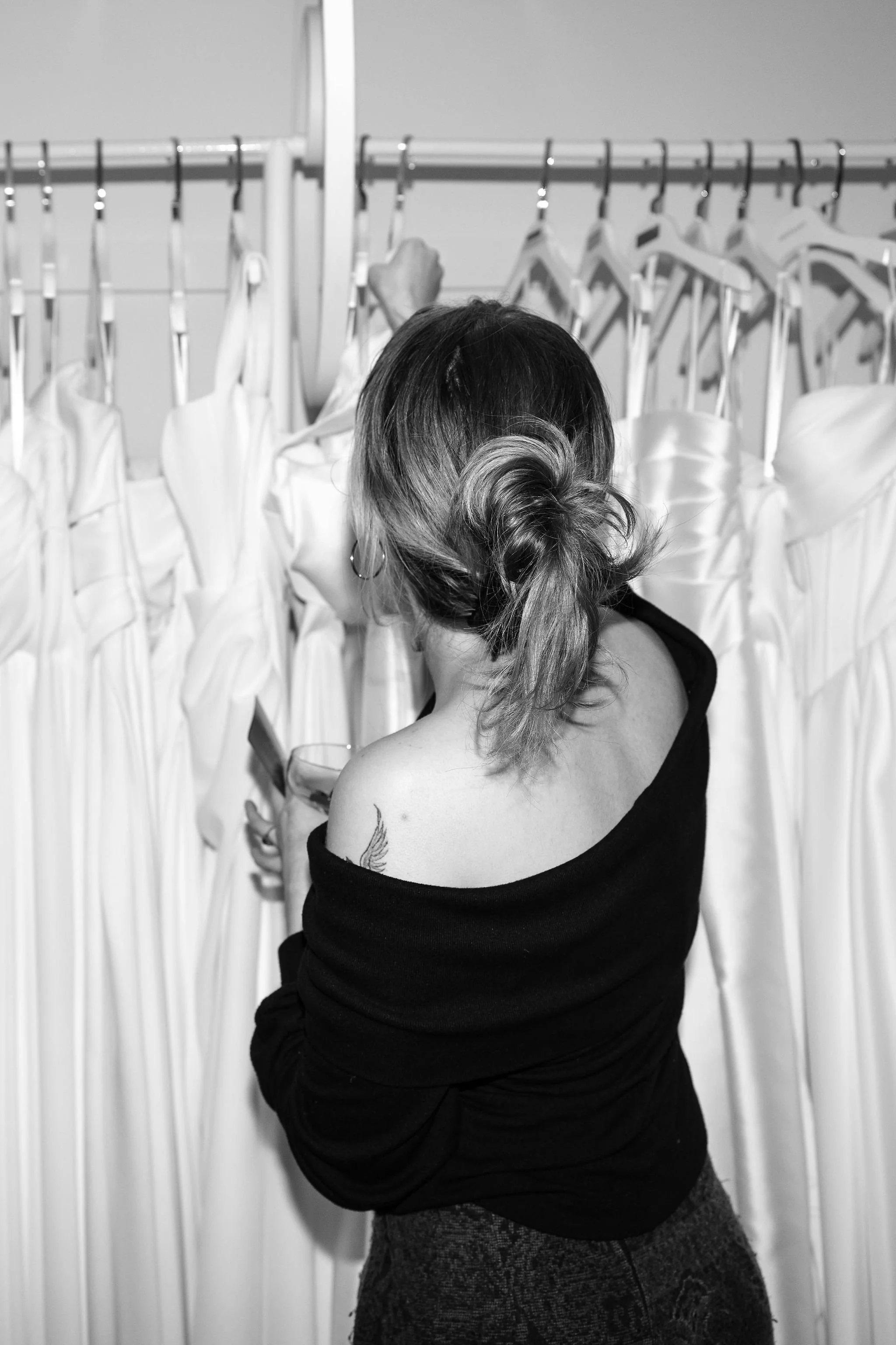 A woman with a tattoo on her shoulder, holding a glass of wine, looking at a row of white dresses on hangers.