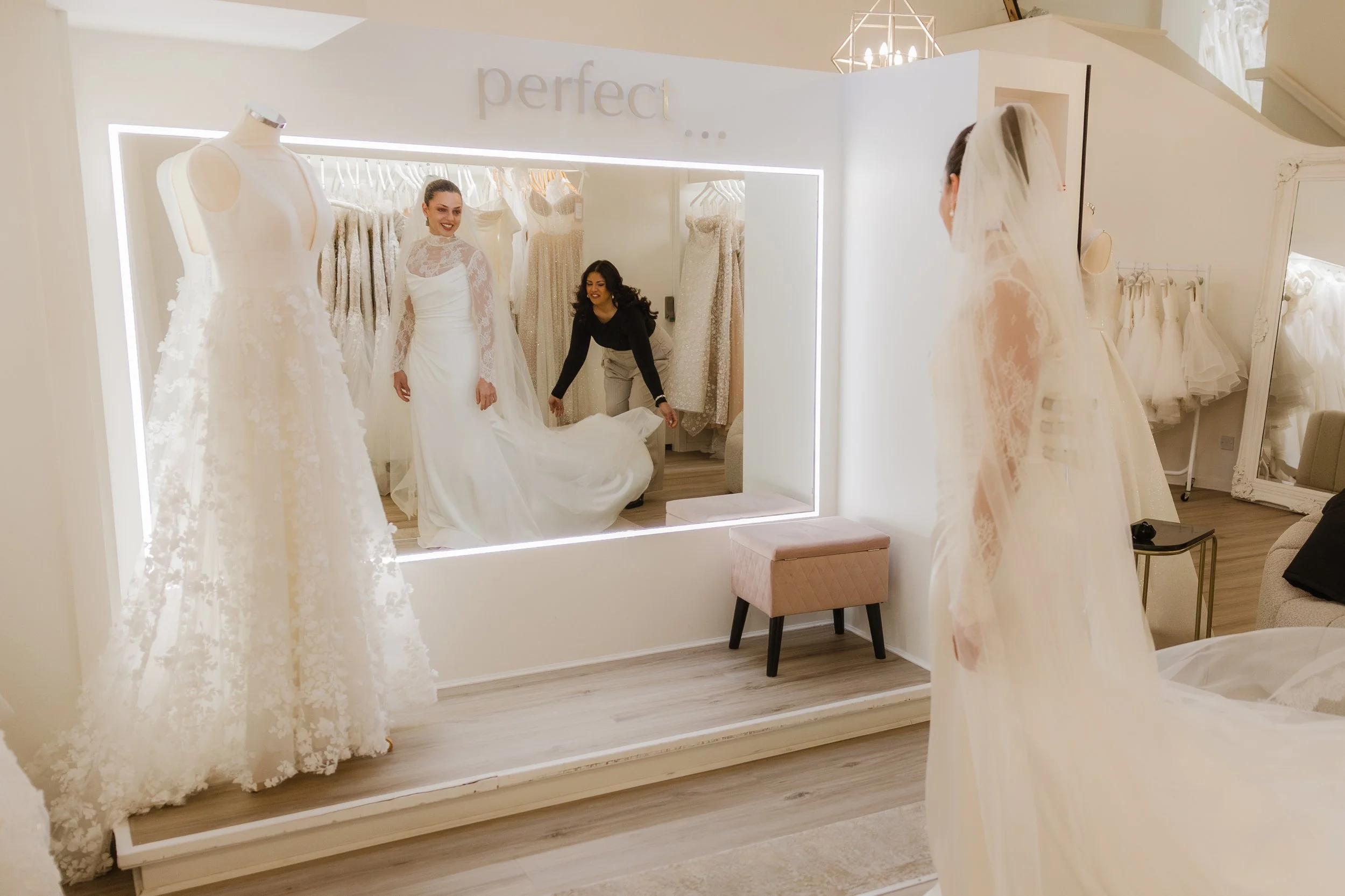 A woman trying on a wedding dress at a bridal boutique with the help of a staff member. The bride is reflected in a large mirror, and several wedding dresses are displayed on mannequins and racks in the background. The boutique has a bright, elegant interior with a sign that reads 'perfect...' above the mirror.