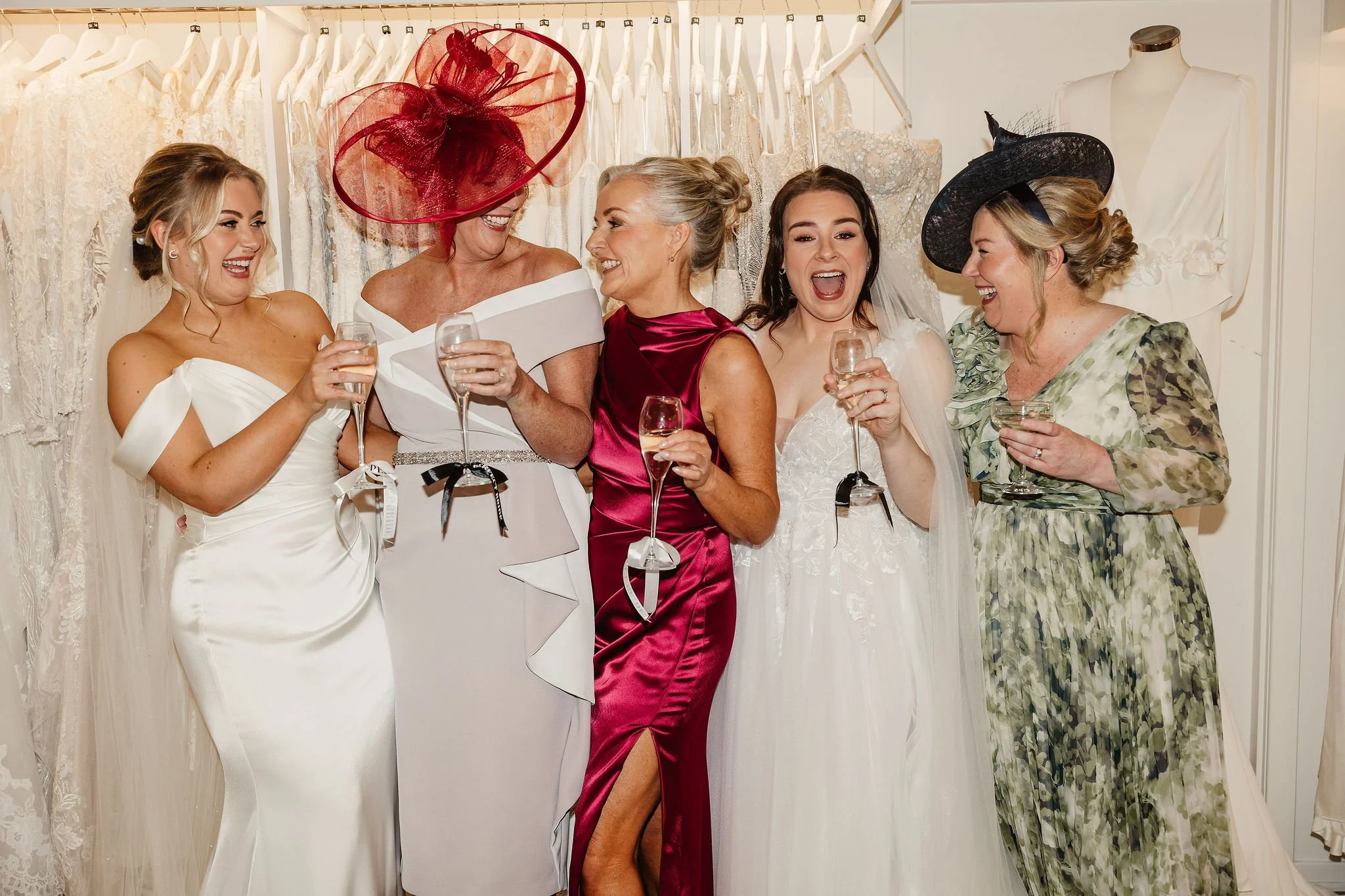 Six women dressed in elegant wedding and formal dresses celebrating and laughing together, holding wine glasses, in a bridal shop or dressing room with wedding gowns hanging on racks in the background.