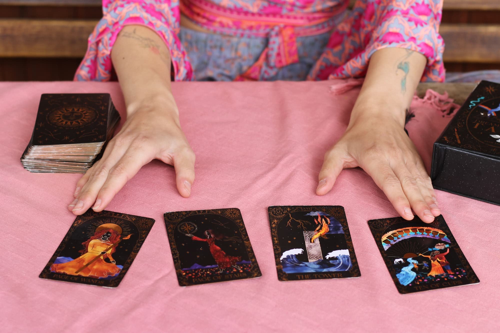 Person with tarot cards spread on pink tablecloth, hands hovering over four tarot cards: Page of Wands, Page of Stars, The Tower, and Ten of Cups.