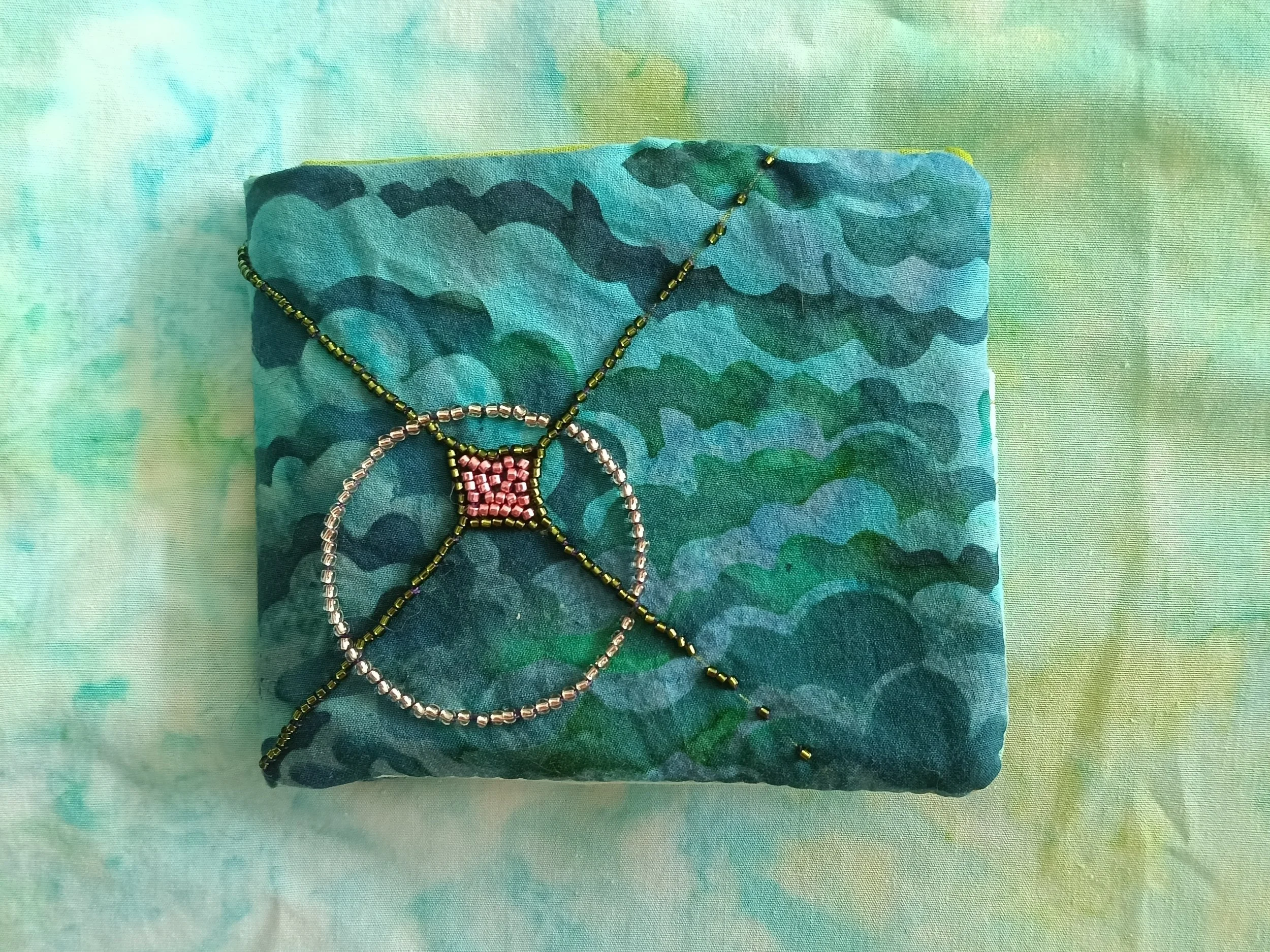 Close-up of a beaded wallet with pink, silver, and gold beads on a blue, green, and teal tie-dye fabric background.