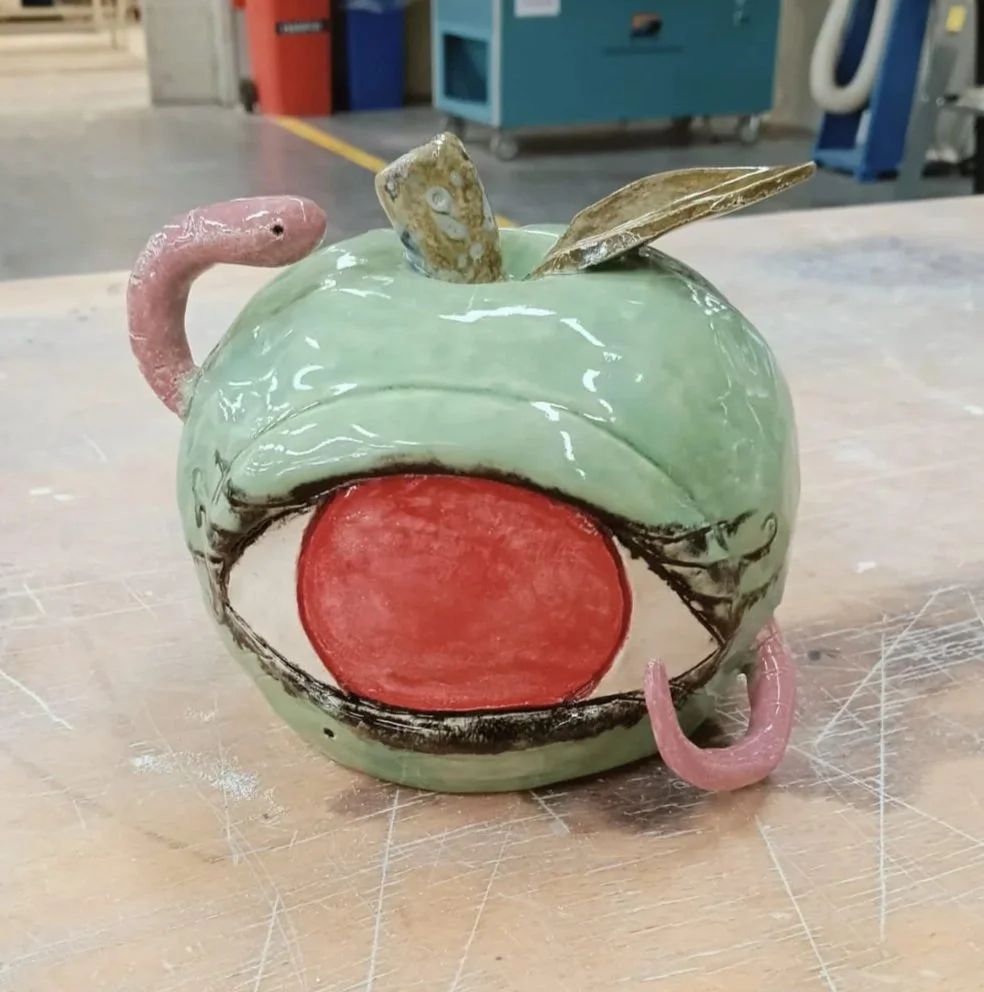 A ceramic teapot designed to look like a cartoon character with large eyes and an open mouth, featuring a pink handle and spout, and a sea shell on top.
