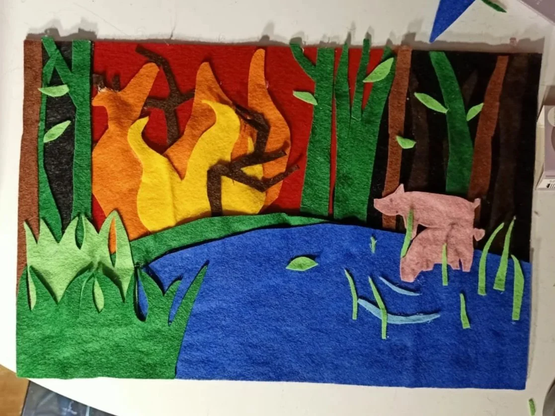 Felt art depicting a forest fire scene with trees, a creek, and a pink pig.