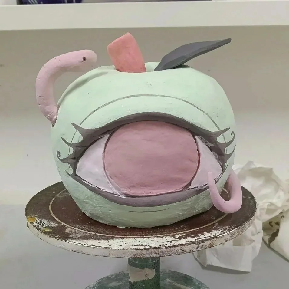 A green teapot with a painted cartoon face, large open mouth, and eye brows, sitting on a rotating stand. The teapot has a pink handle and a gray lid, with a pink tongue and a gray spoon on top.