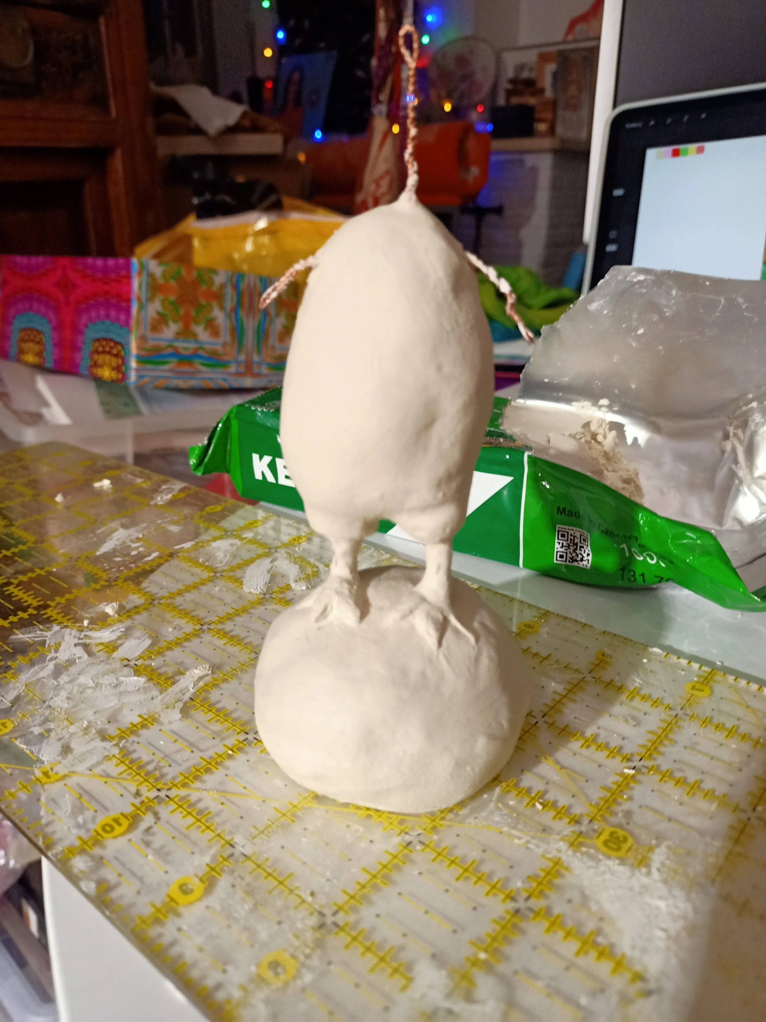 A sculpture of a bird with a rounded body, standing on a circular base, made from a white or off-white material, possibly clay or plaster, on a table with a yellow grid pattern, surrounded by a partially opened plastic bag and a green package. In the