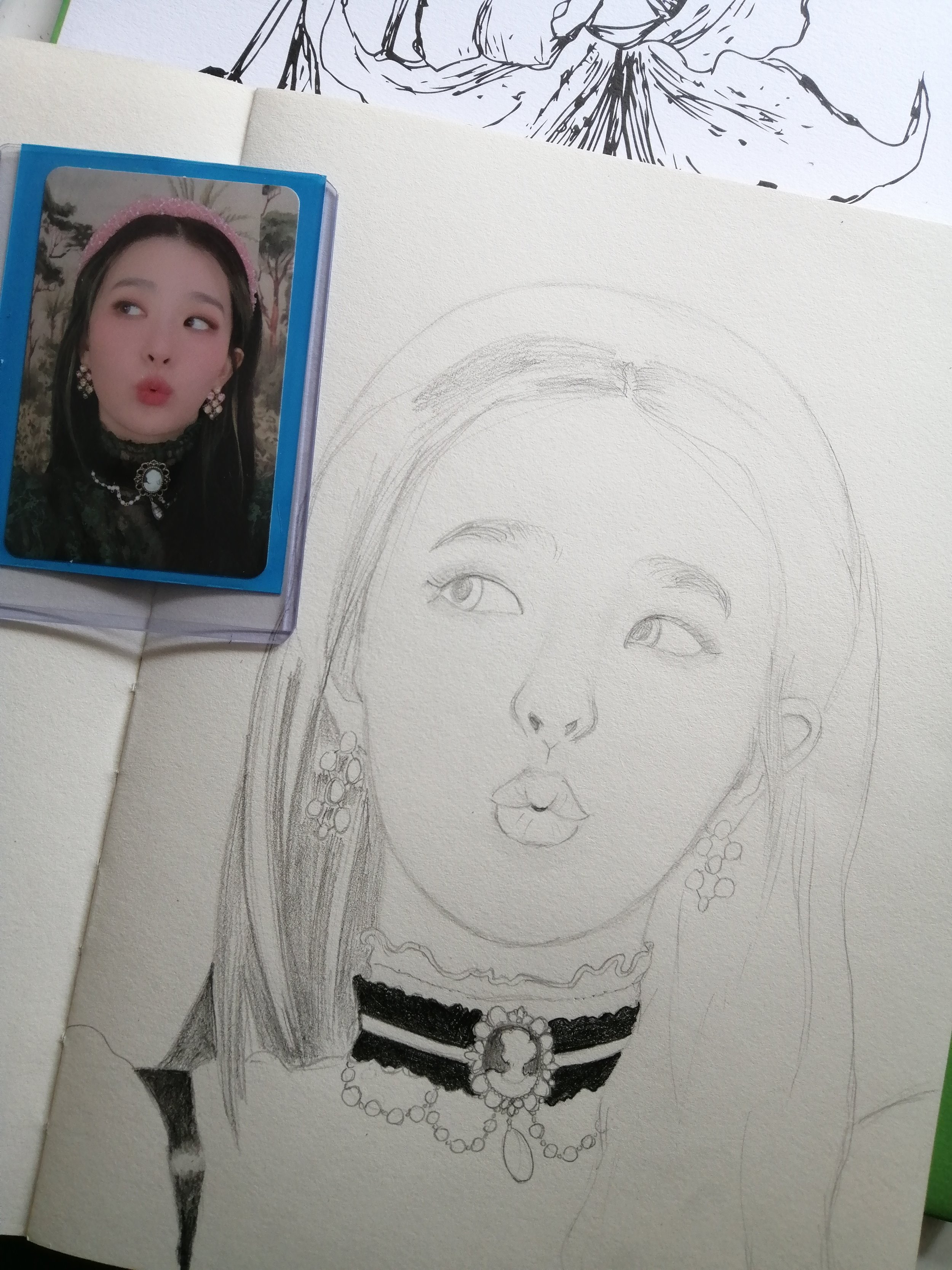 A pencil sketch of a woman's face on a piece of paper with a reference photo of a woman wearing jewelry and a pink headband, displayed nearby. The woman has long hair, earrings, a necklace, and a necklace with a large pendant.