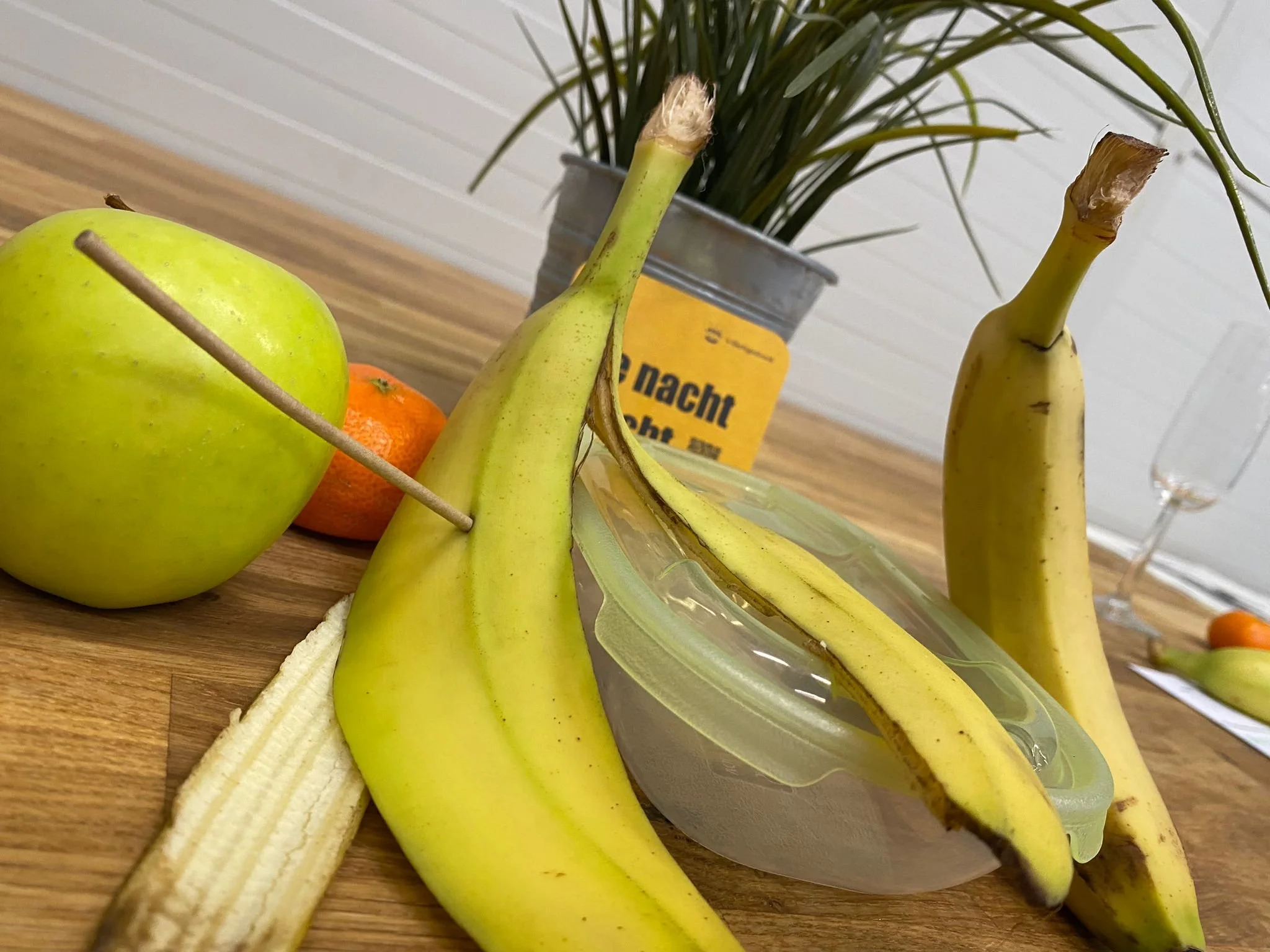 Fruits including a green apple, banana, and an orange with a banana peel, with a potted plant and a yellow container in the background.