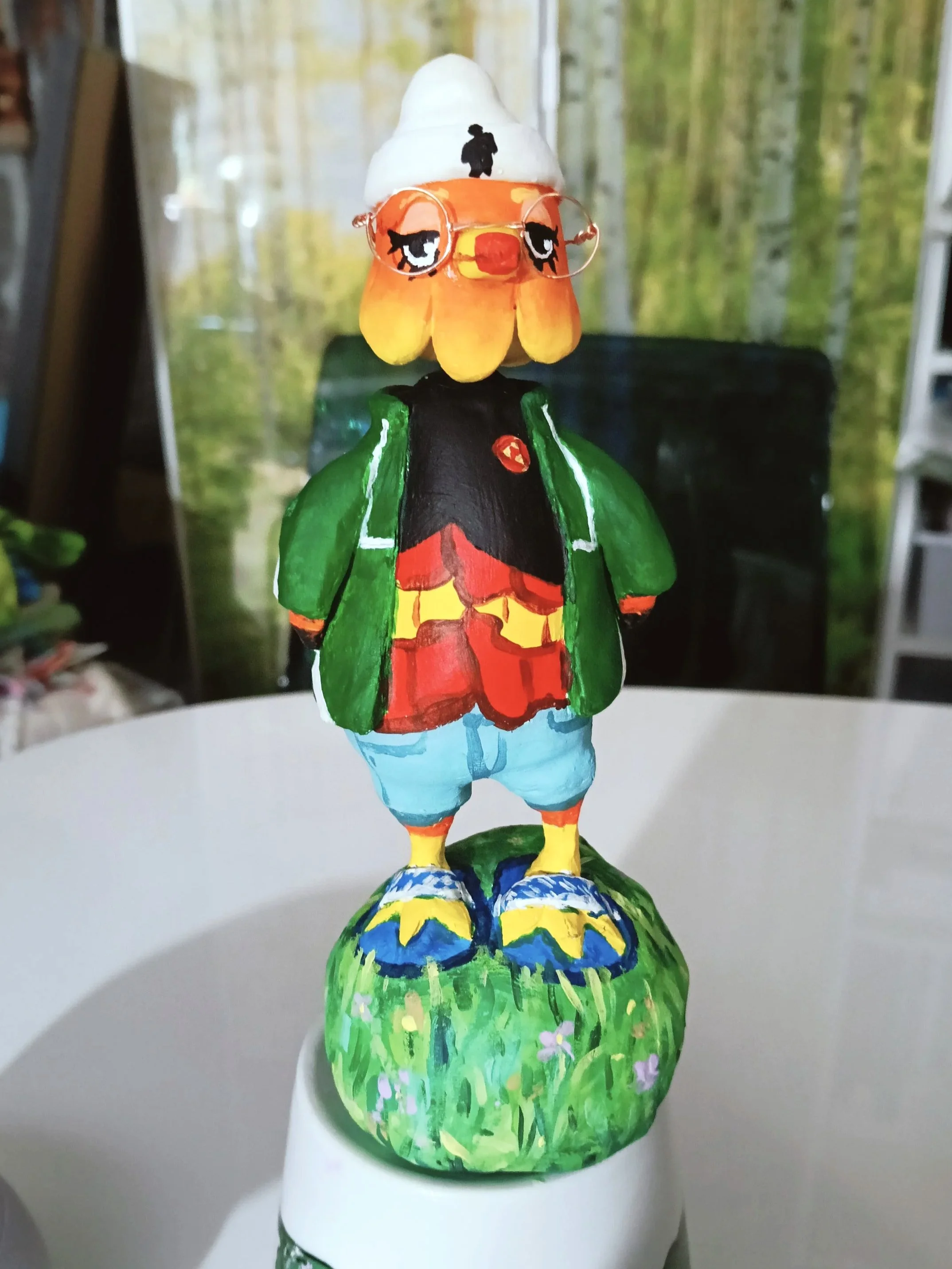 Colorfully painted bird figurine standing on a green base with flowers, wearing blue shoes, red shorts, black shirt, green jacket, and a white hat with a small figure silhouette on top.