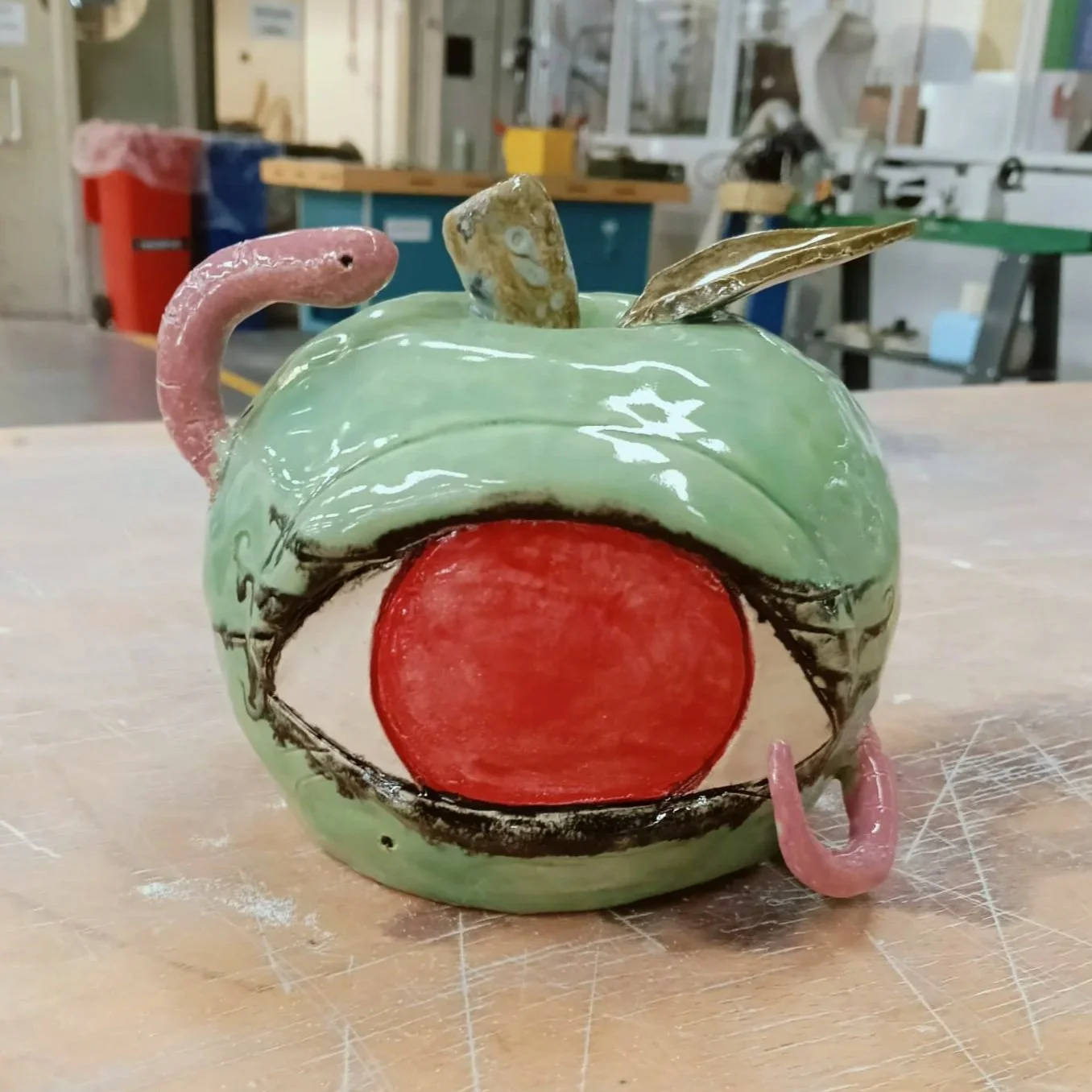 A ceramic sculpture of a cartoonish face with an open mouth, green face, red tongue, and pink tentacle-like features on top and sides, placed on a wooden surface in a workshop.