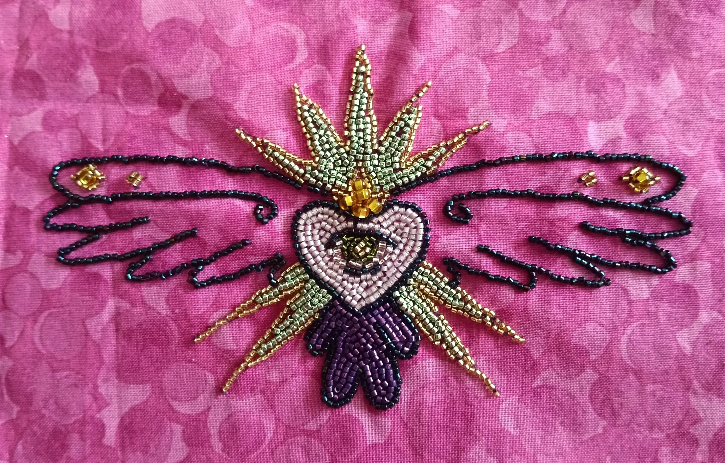 Beaded embroidery of a fairy with purple wings and a golden crown on pink fabric.